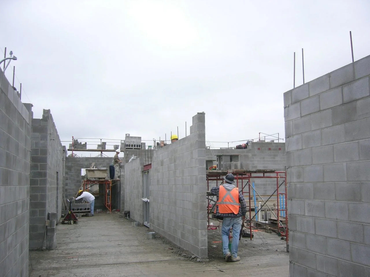 Construction workers building walls with concrete blocks on a cloudy day, some workers on scaffolding and others walking through the site.
