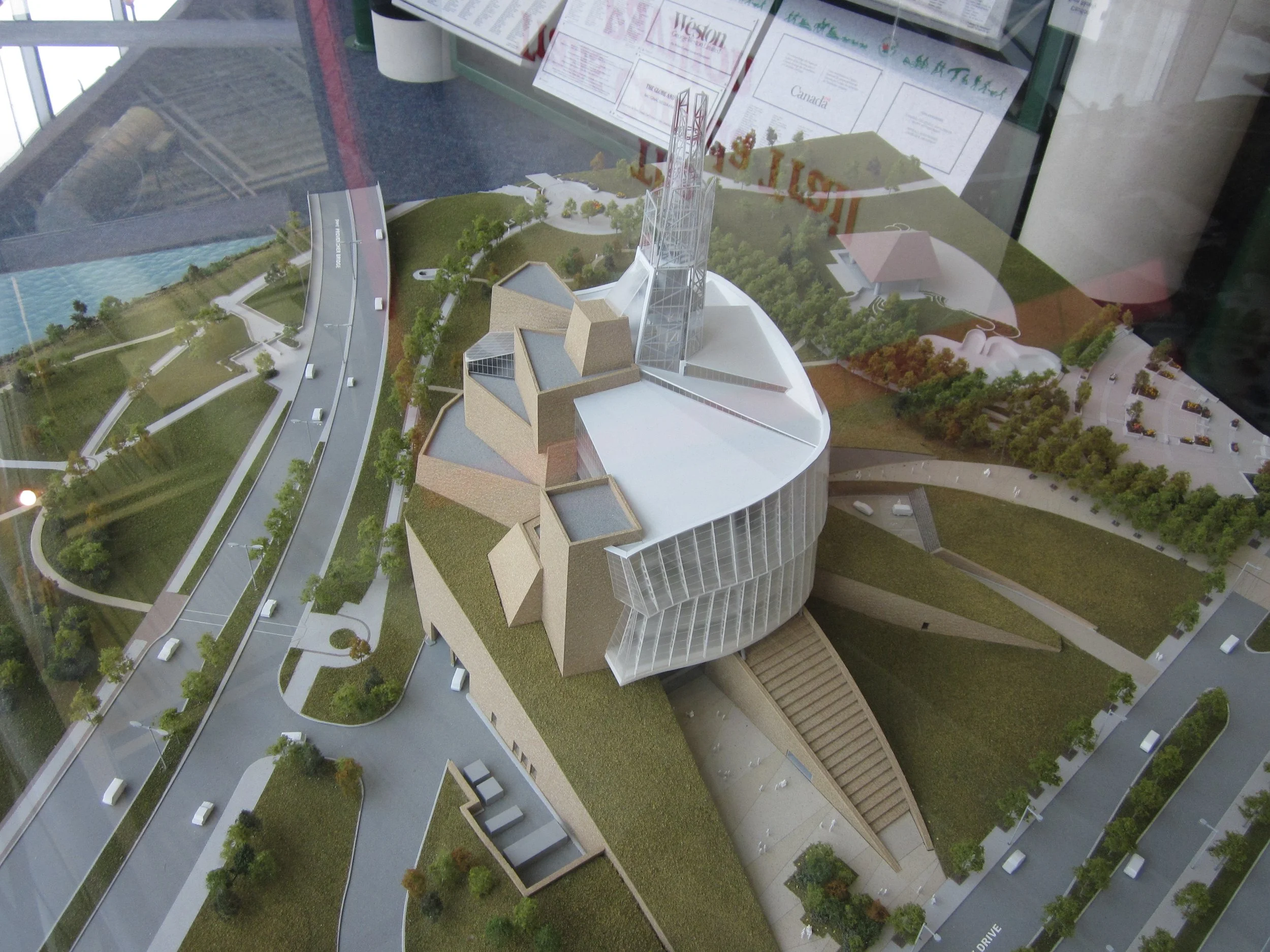 A detailed architectural scale model of a modern building with various geometric features, green roofs, and surrounding landscape, viewed through a glass case.