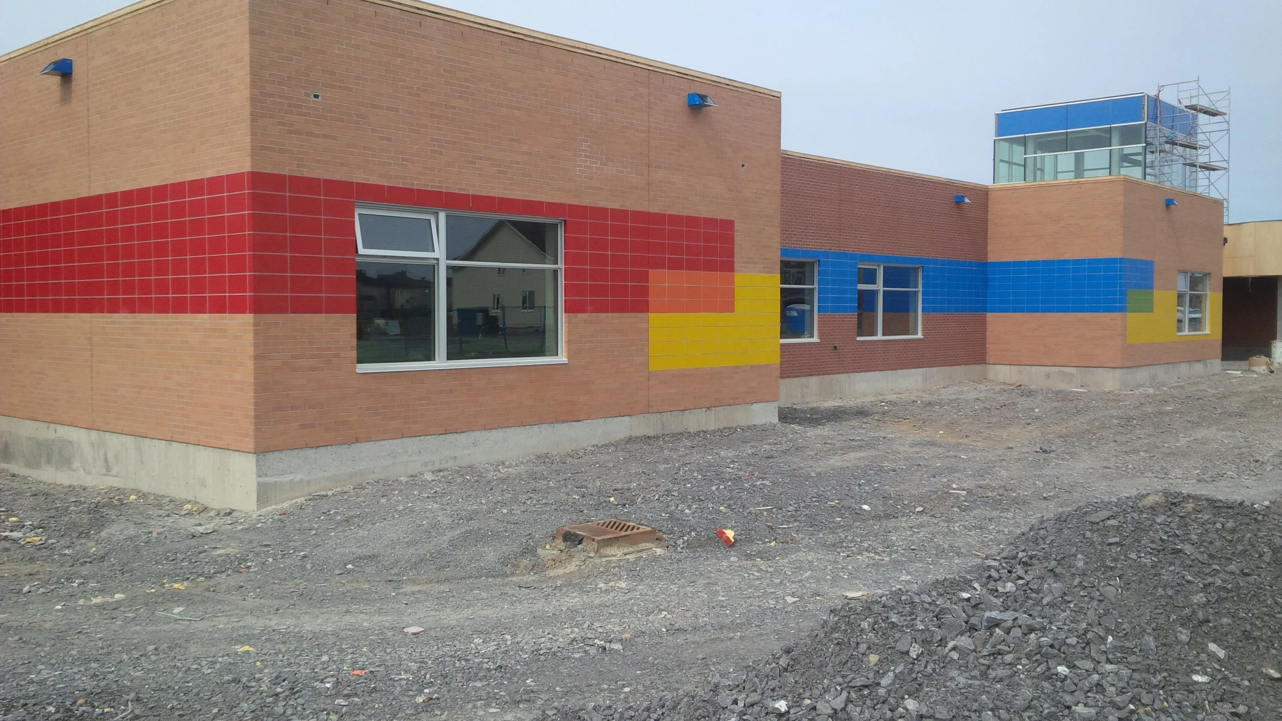 Building under construction with colorful geometric tile patterns in red, yellow, and blue on its brick exterior, construction materials and dirt in the foreground.