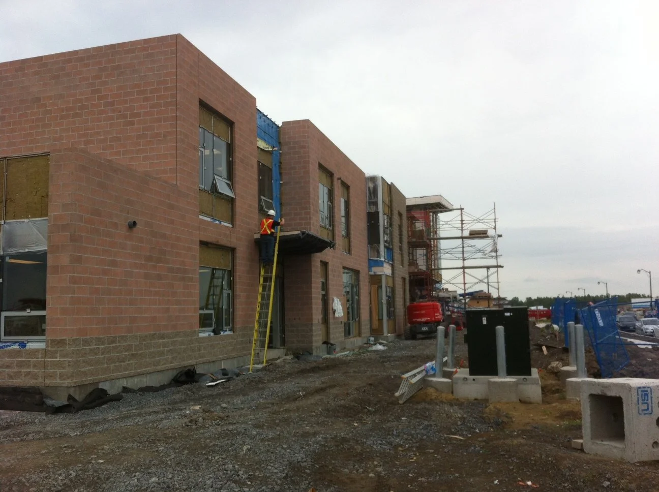 Construction site with workers installing windows on a brick building, scaffolding on the right, and construction materials on the ground.