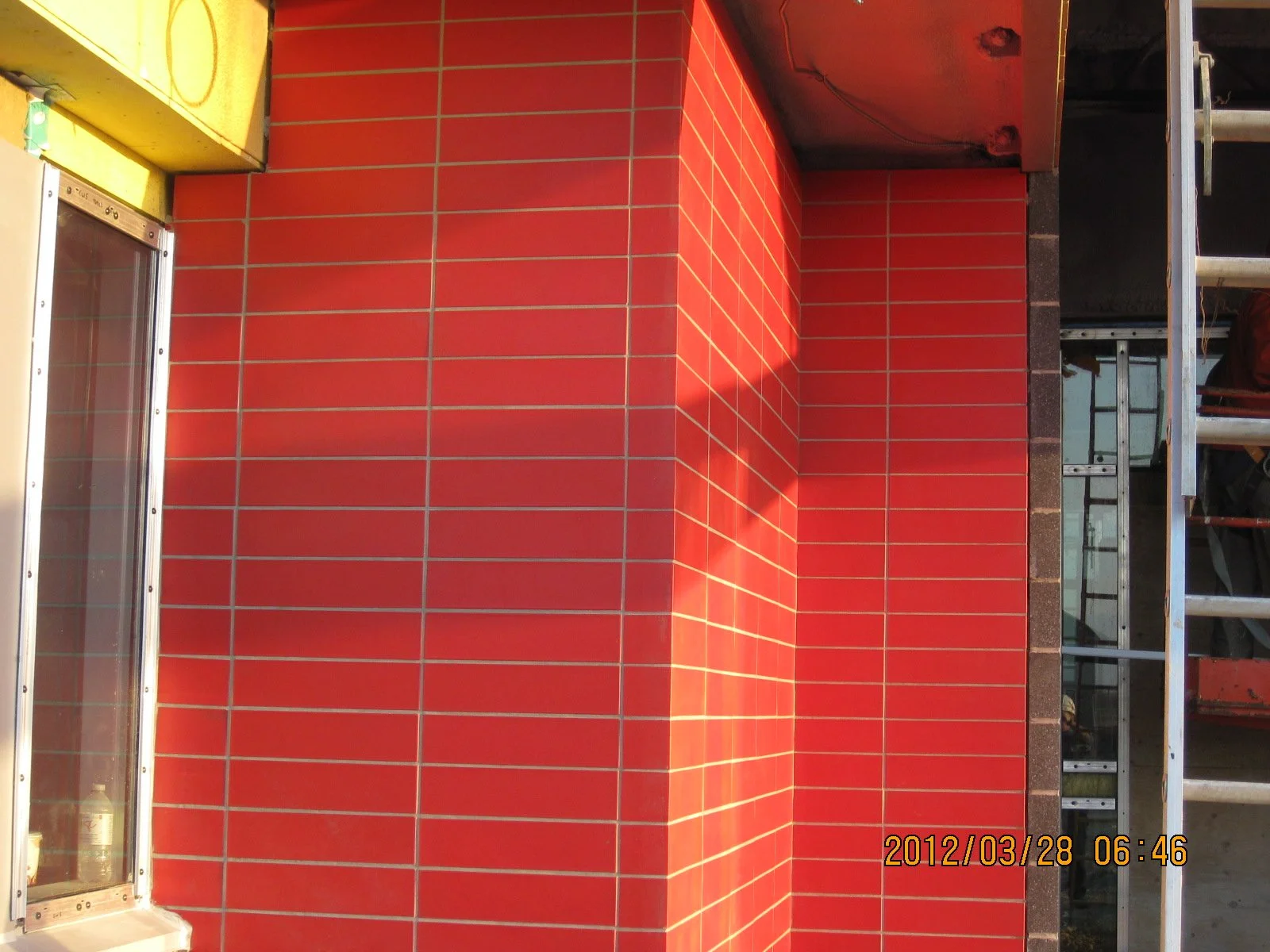 Image showing a corner of a building with red tiled exterior walls, a glass display case on the left, and a ladder on the right. The photo is taken outdoors in sunlight. The timestamp on the photo indicates it was taken on March 28, 2012, at 6:46.