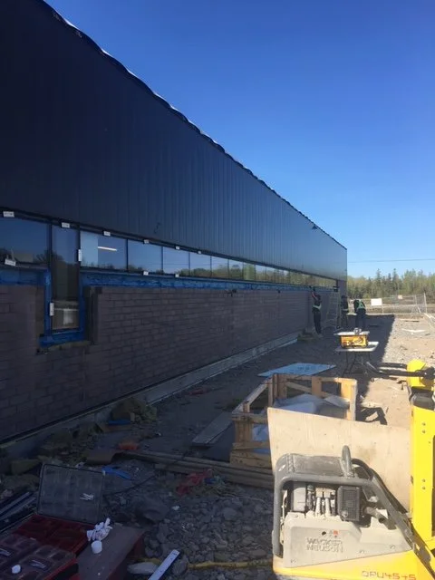 Construction site with a long building, glass windows, and workers in the background, under a clear blue sky.