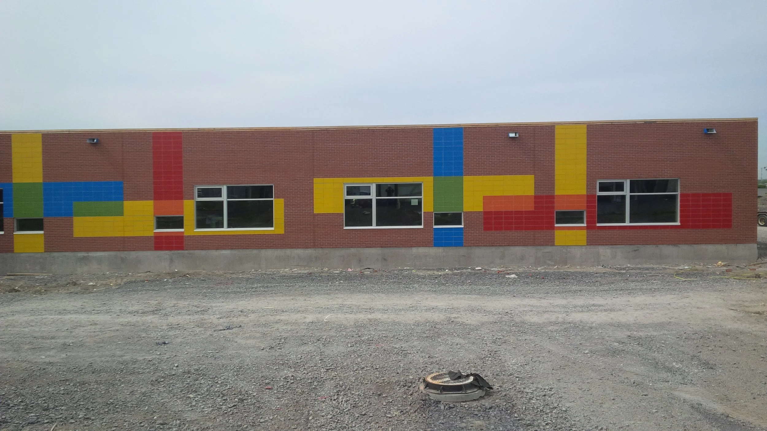 Brick building with colorful geometric mural, windows, and four exterior lights on a cloudy day, surrounded by dirt ground.