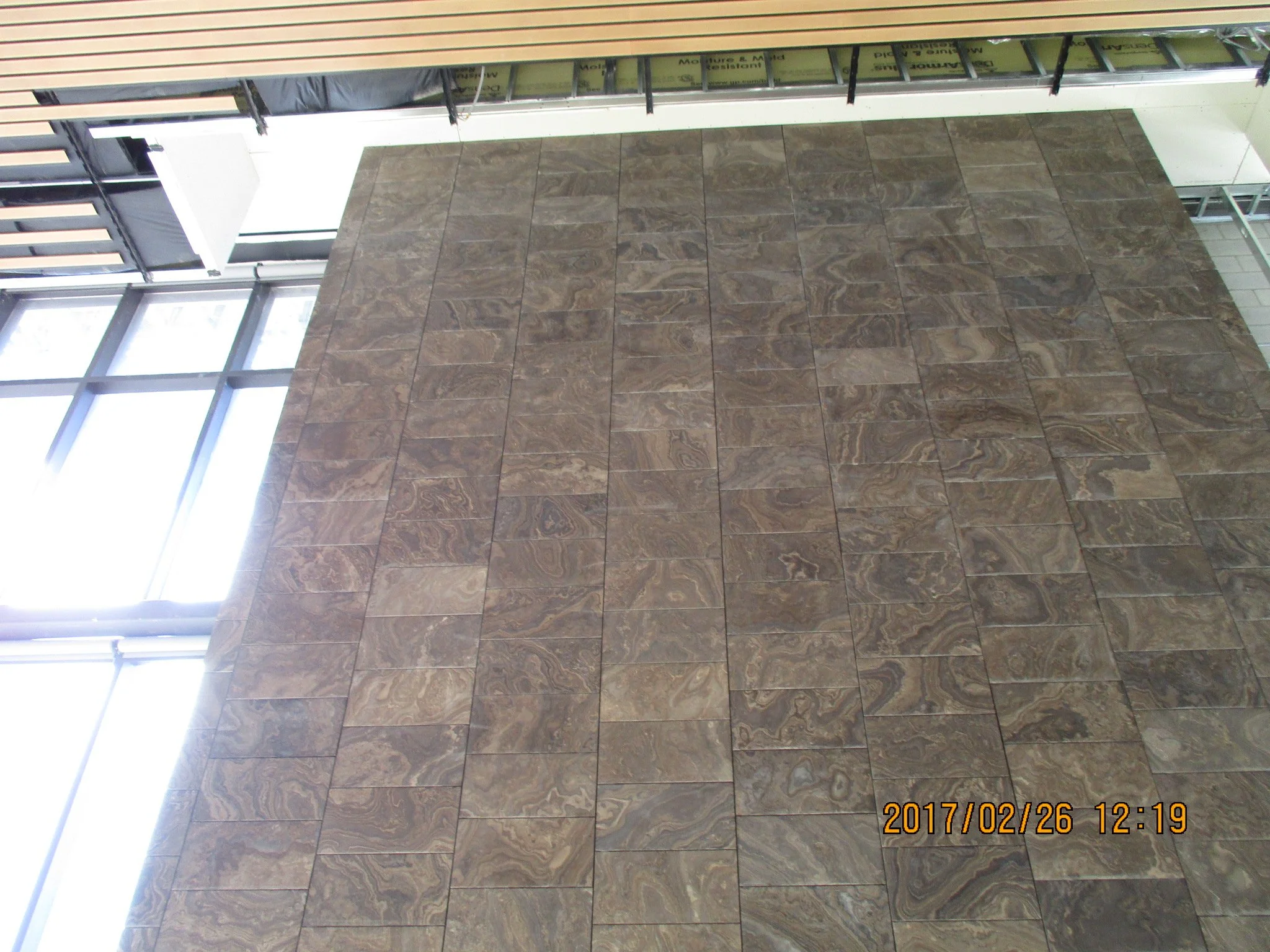 Interior view of a building under construction, showing a wall with brown tiled stone pattern and large glass windows.