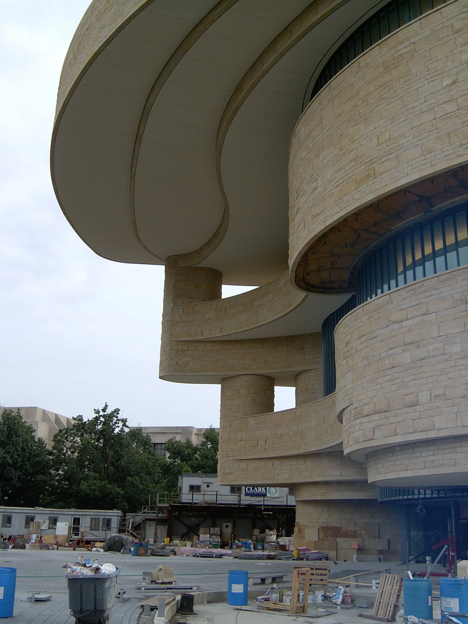 Construction site with a modern, curved building made of beige stone, featuring rounded balconies and large overhanging roof sections.