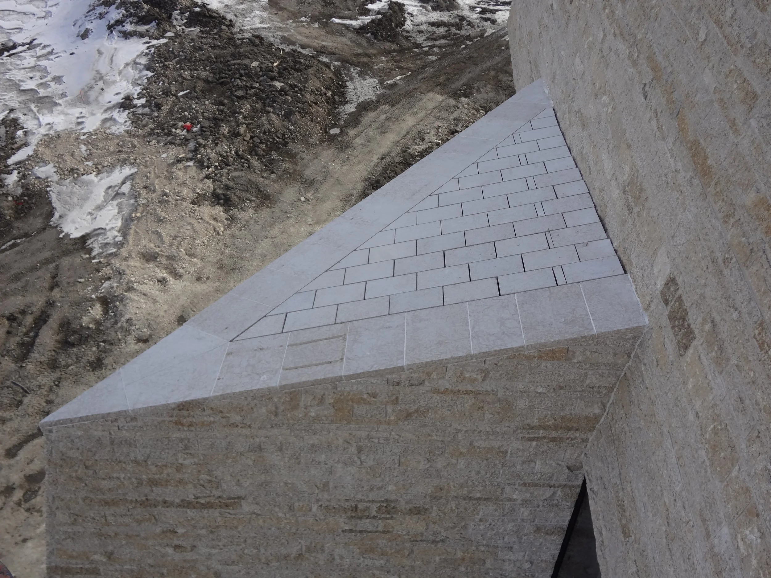 Close-up of a corner stone wall with brickwork pattern, adjacent to a concrete step, in a construction site with dirt and snow in the background.
