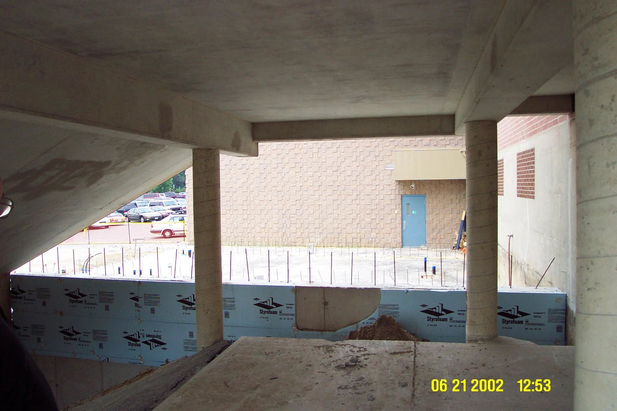 View from inside a building under construction, showing concrete columns, partial walls with Styrofoam insulation, and a construction site outside with parked cars and a brick wall.