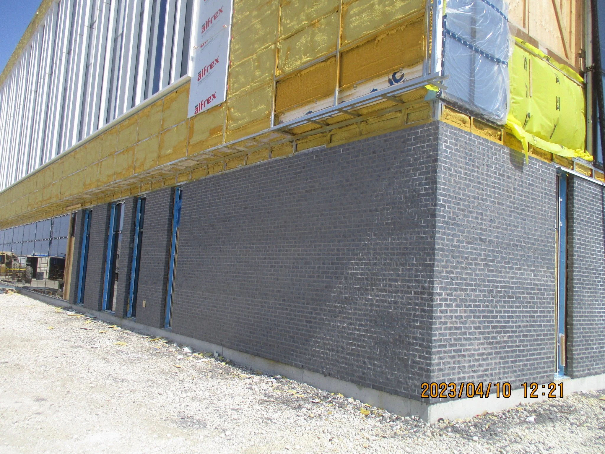Construction site showing a building with a black brick exterior wall and yellow insulation on the upper level, with windows and construction materials around.