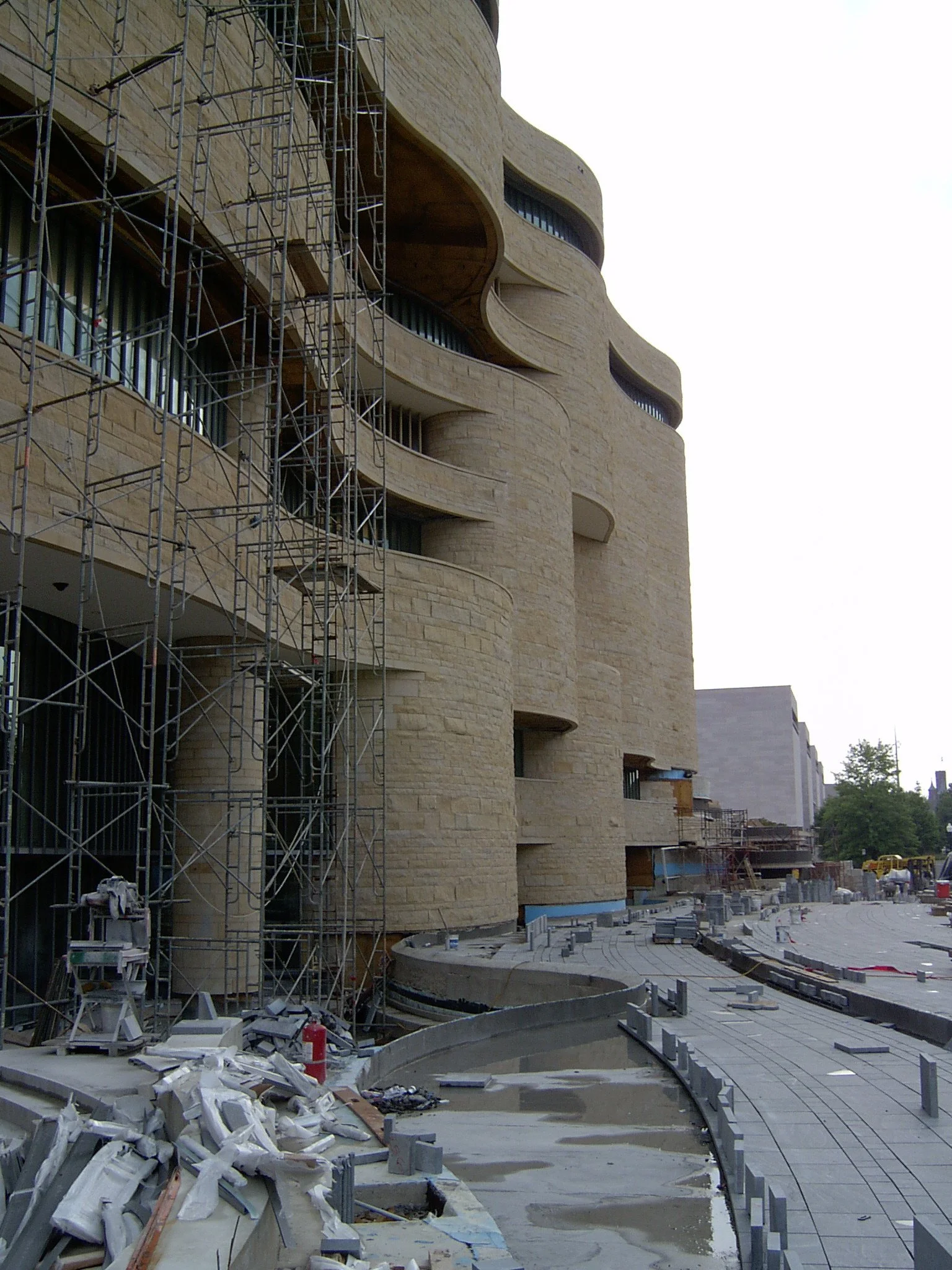 Building under construction with scaffolding and a curved stone facade, paving work in progress on a sidewalk.