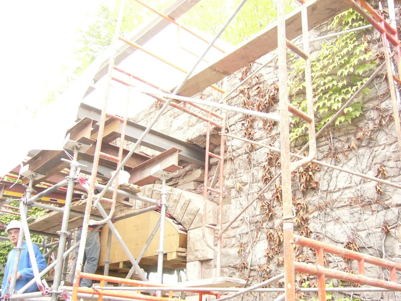 Construction site with scaffolding in front of a stone wall covered with ivy and dried leaves, two workers wearing helmets are visible at the lower left corner.
