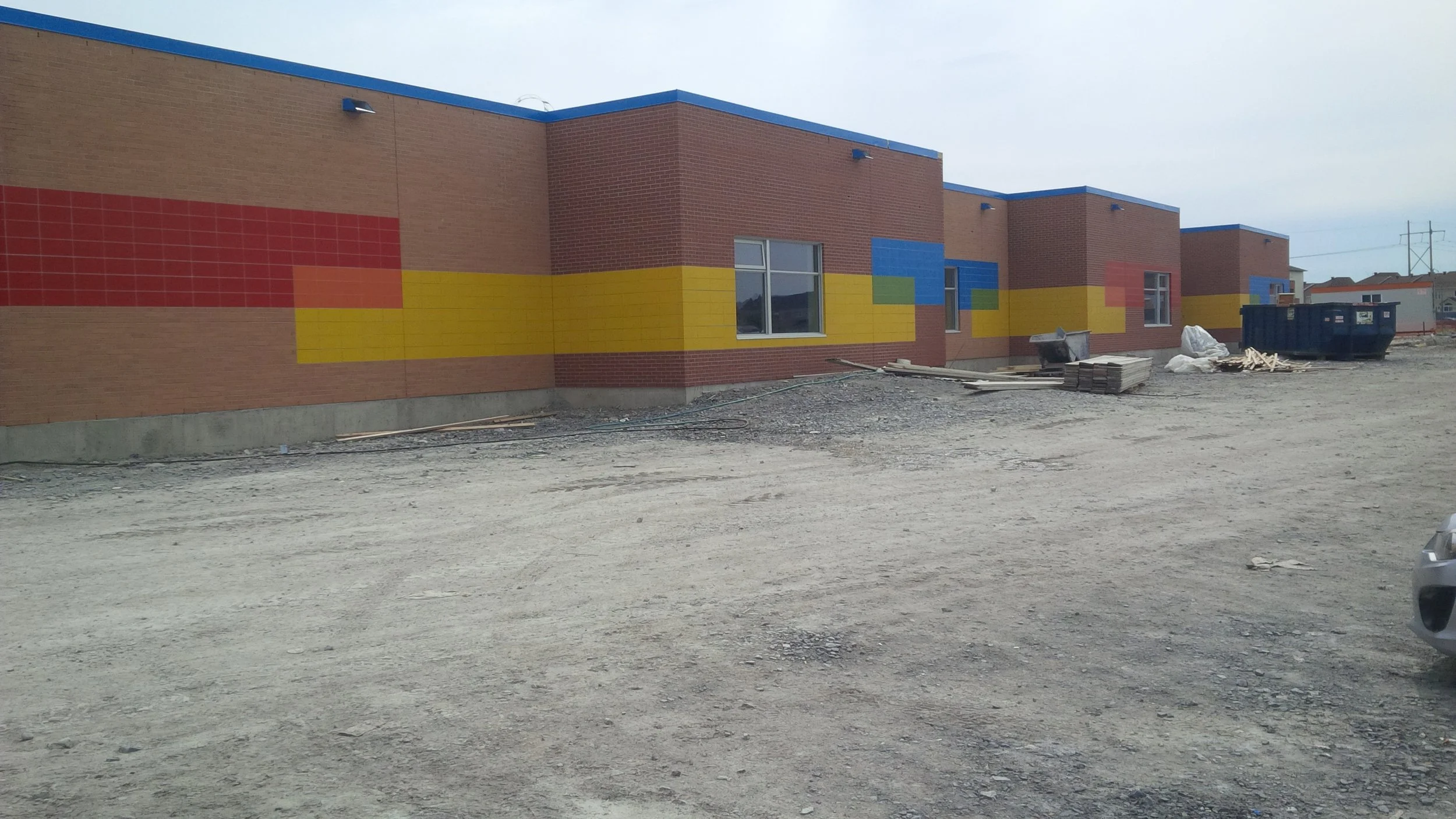A school building under construction with red brick walls and colorful geometric murals, dirt ground in the foreground, and a gray sky overhead.