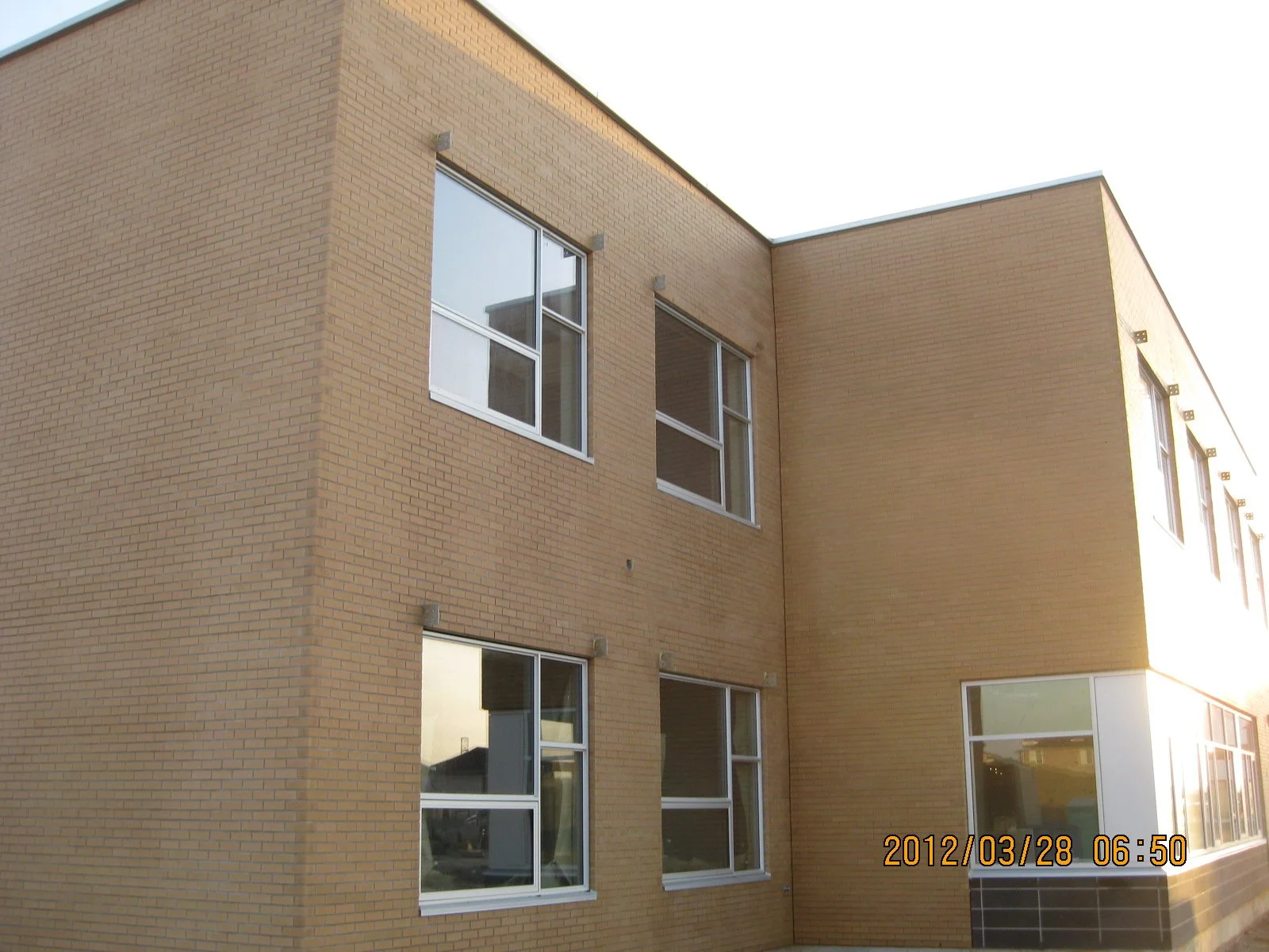 A brick building with multiple windows, some reflecting neighboring structures, and a timestamp indicating it was taken on March 28, 2012, at 6:50 am.