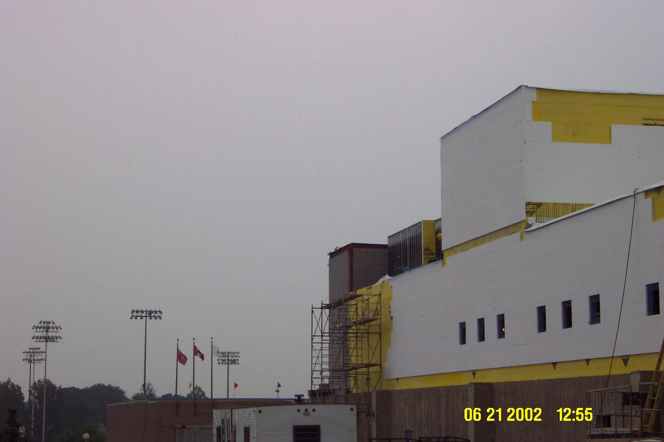 Construction site with a partially built white and yellow building, scaffolding on the side, and flags on tall poles in the background, under a gray cloudy sky.