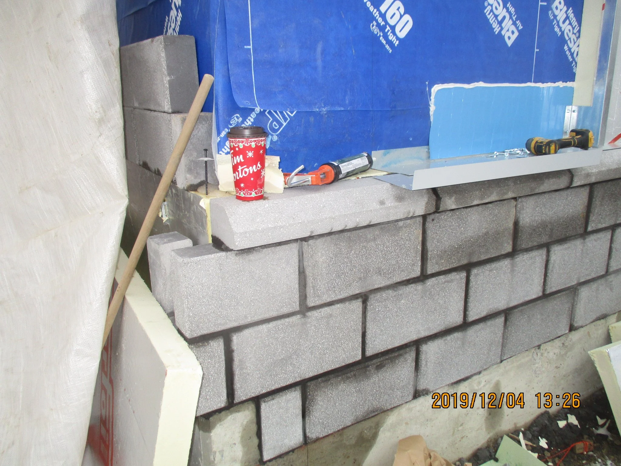 Construction site shows a partially built concrete block wall with tools and materials, including a power drill, caulking gun, a Tim Hortons cup, and a blue insulation sheet.