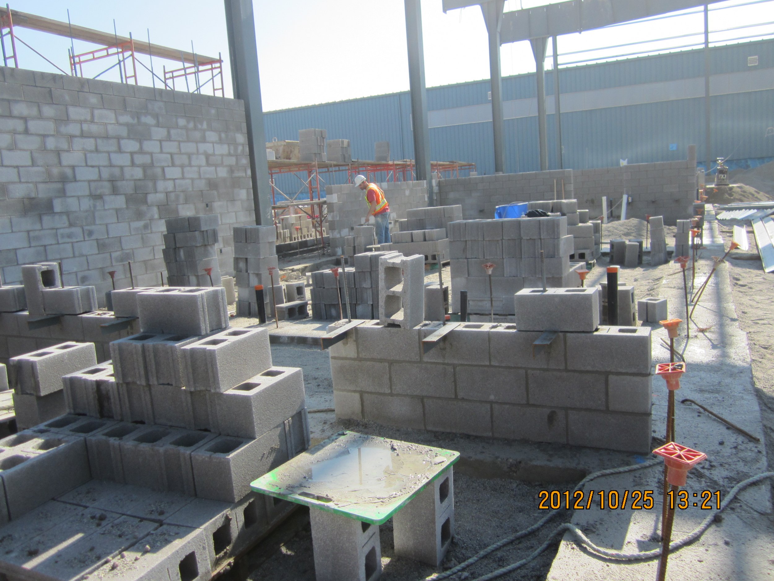 Construction workers building a brick wall at a construction site with stacks of cinder blocks, scaffolding, and construction tools visible.