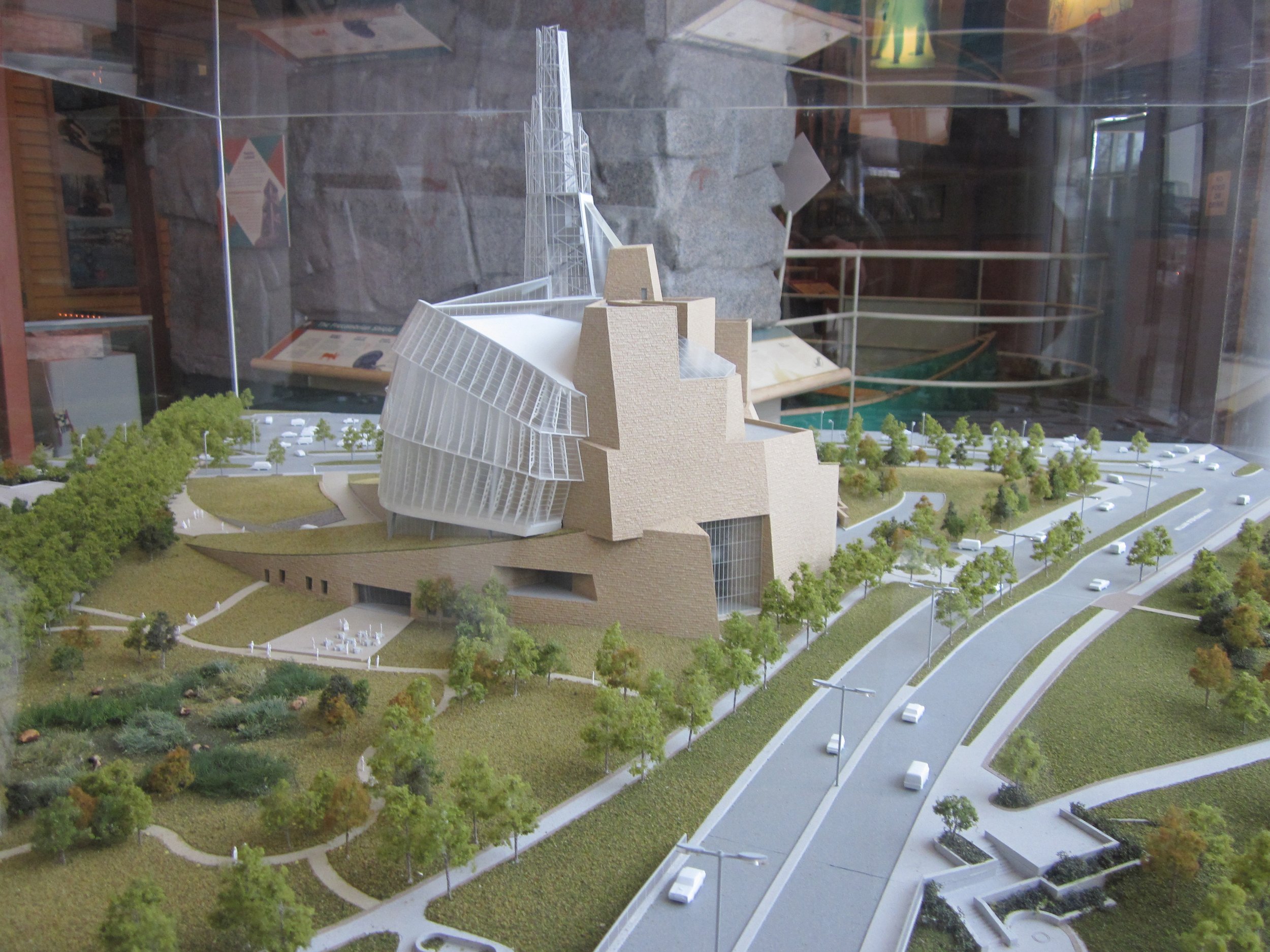 Architectural model of a modern building surrounded by green landscape, trees, pathways, and roads with cars, displayed inside a glass case.