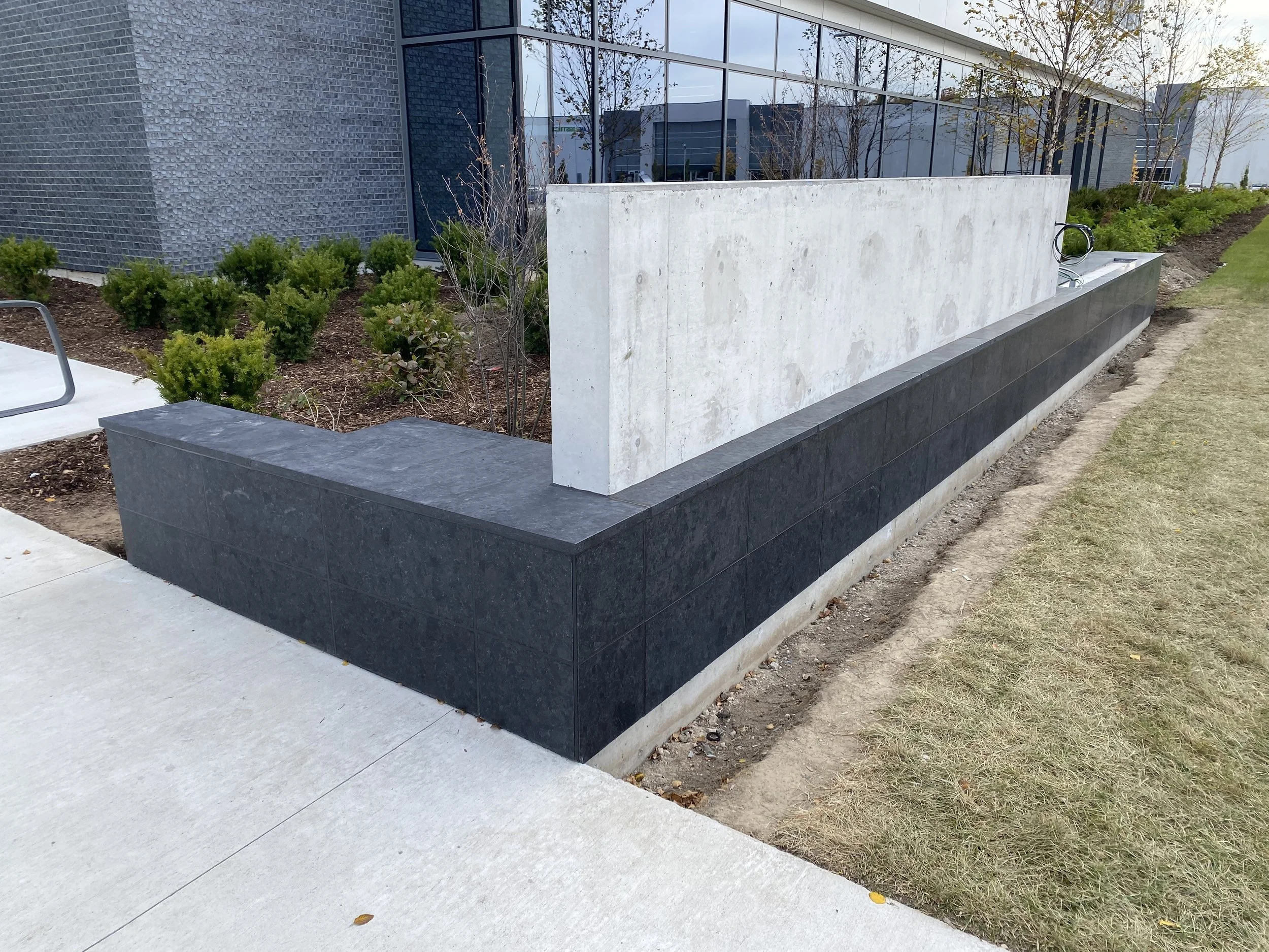 Modern outdoor planter with dark stone base, light concrete panel, and a surrounding landscaped garden, in front of a building with large glass windows.