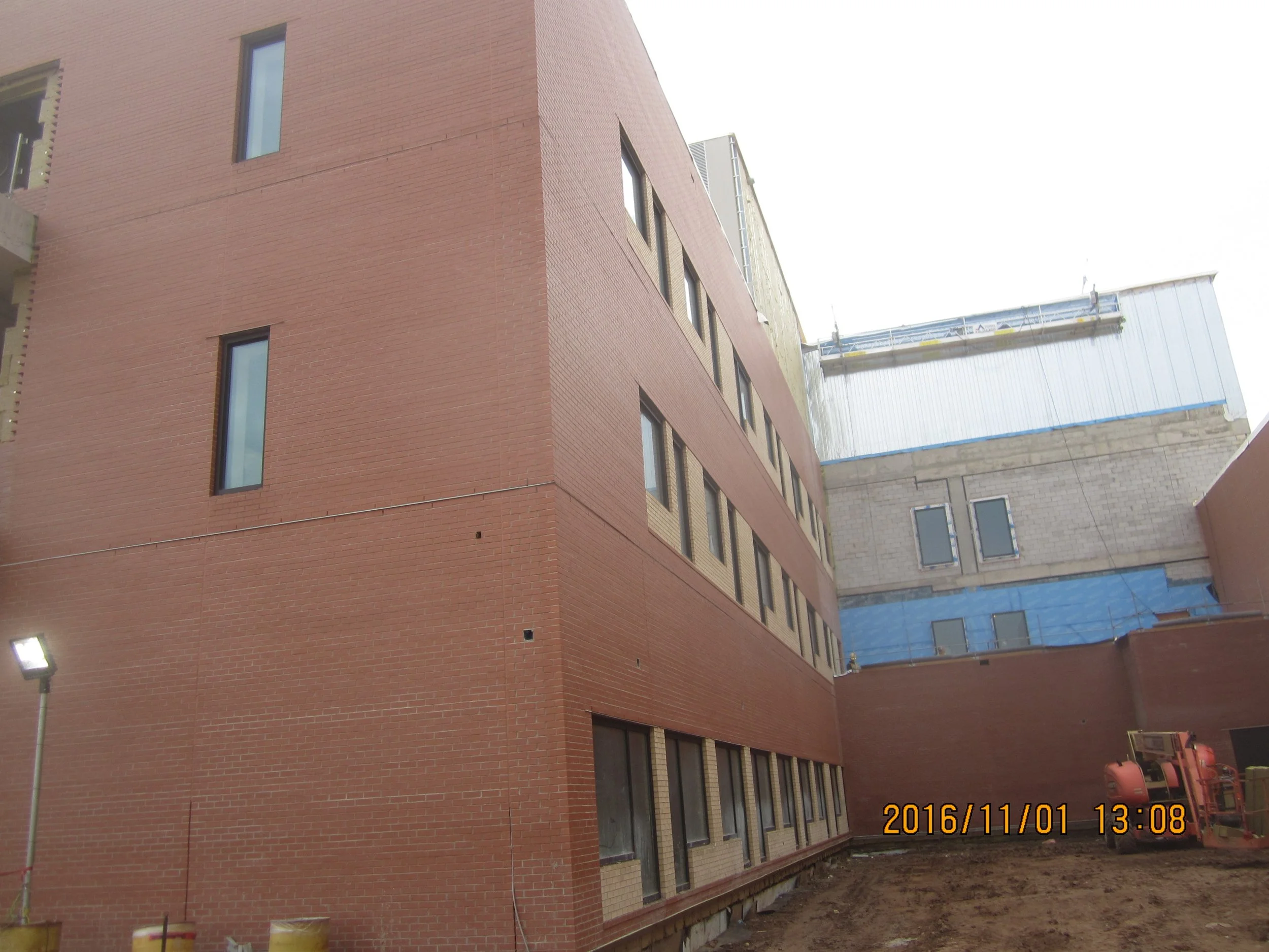 A large red brick building under construction with multiple windows, some sections covered with blue insulation, and construction equipment on the ground.