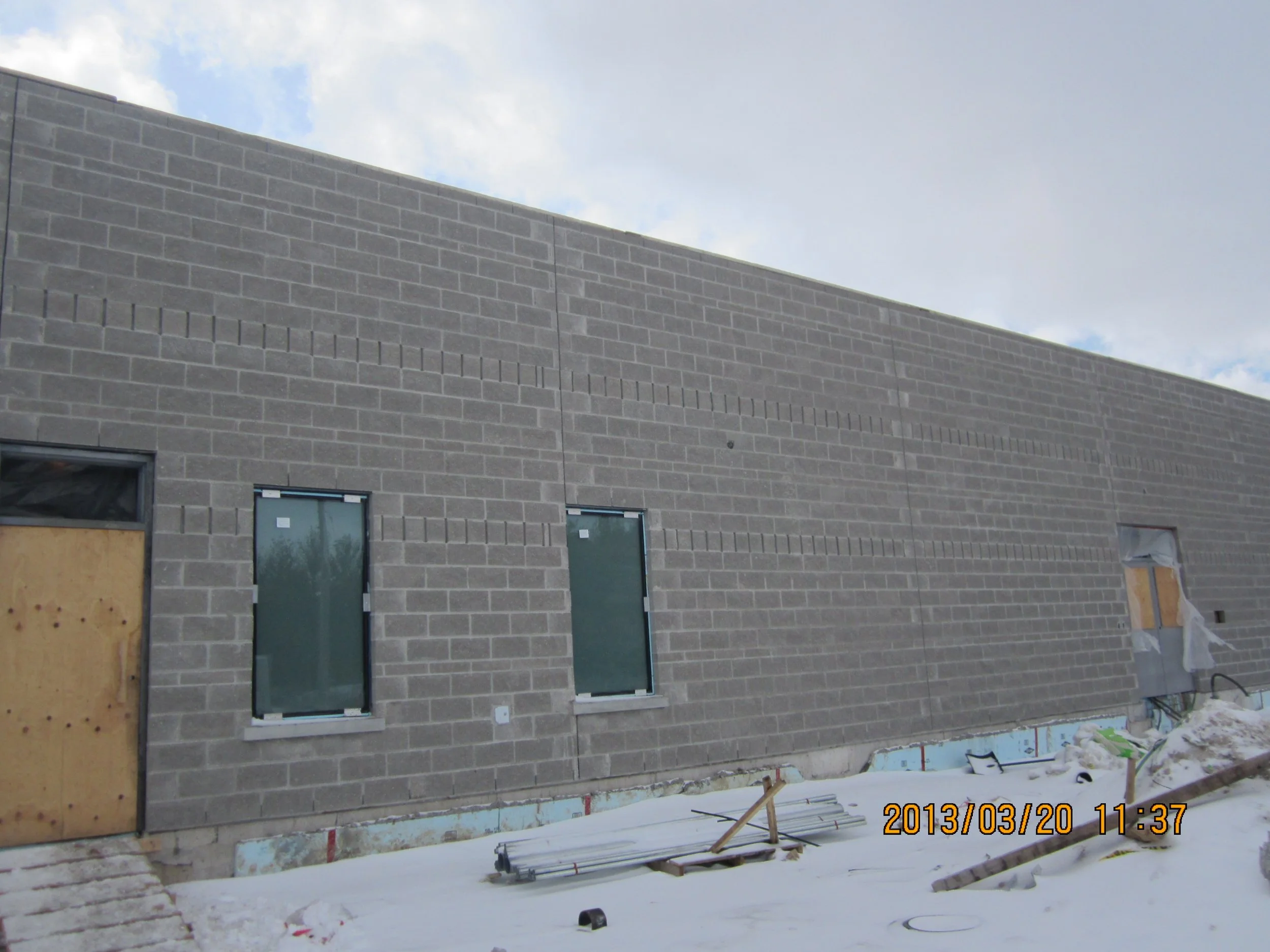 A construction site with a building in progress, showing gray brick walls, two windows, one door boarded up, and snow on the ground.
