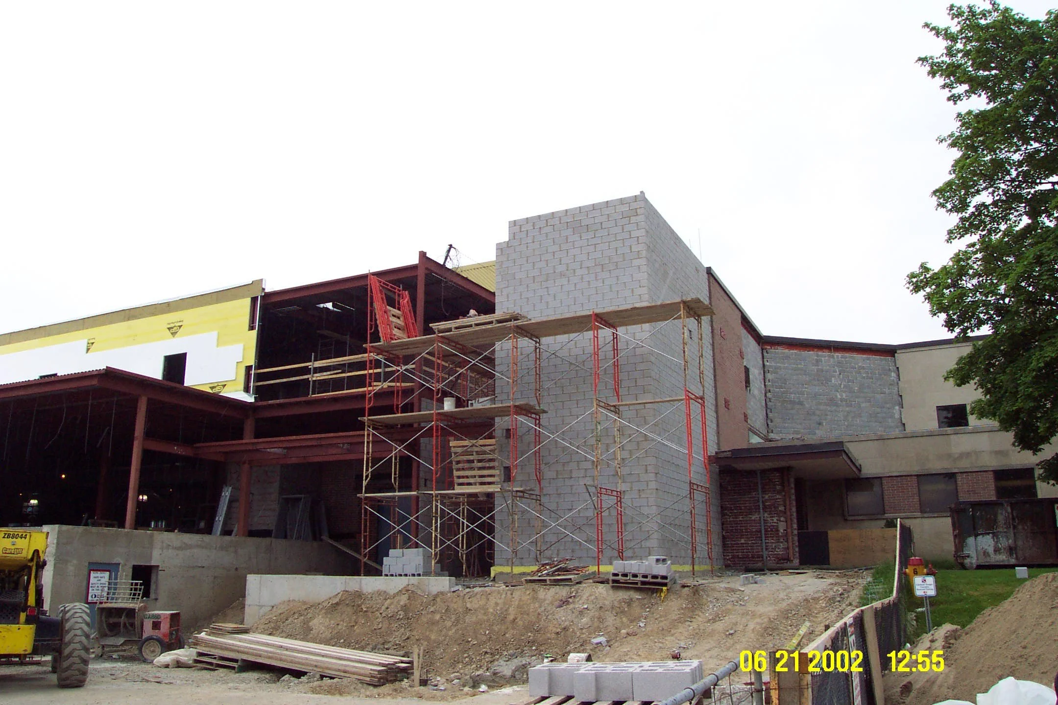 Construction site of a building with scaffolding and partially completed walls, surrounded by dirt and construction equipment, under an overcast sky.