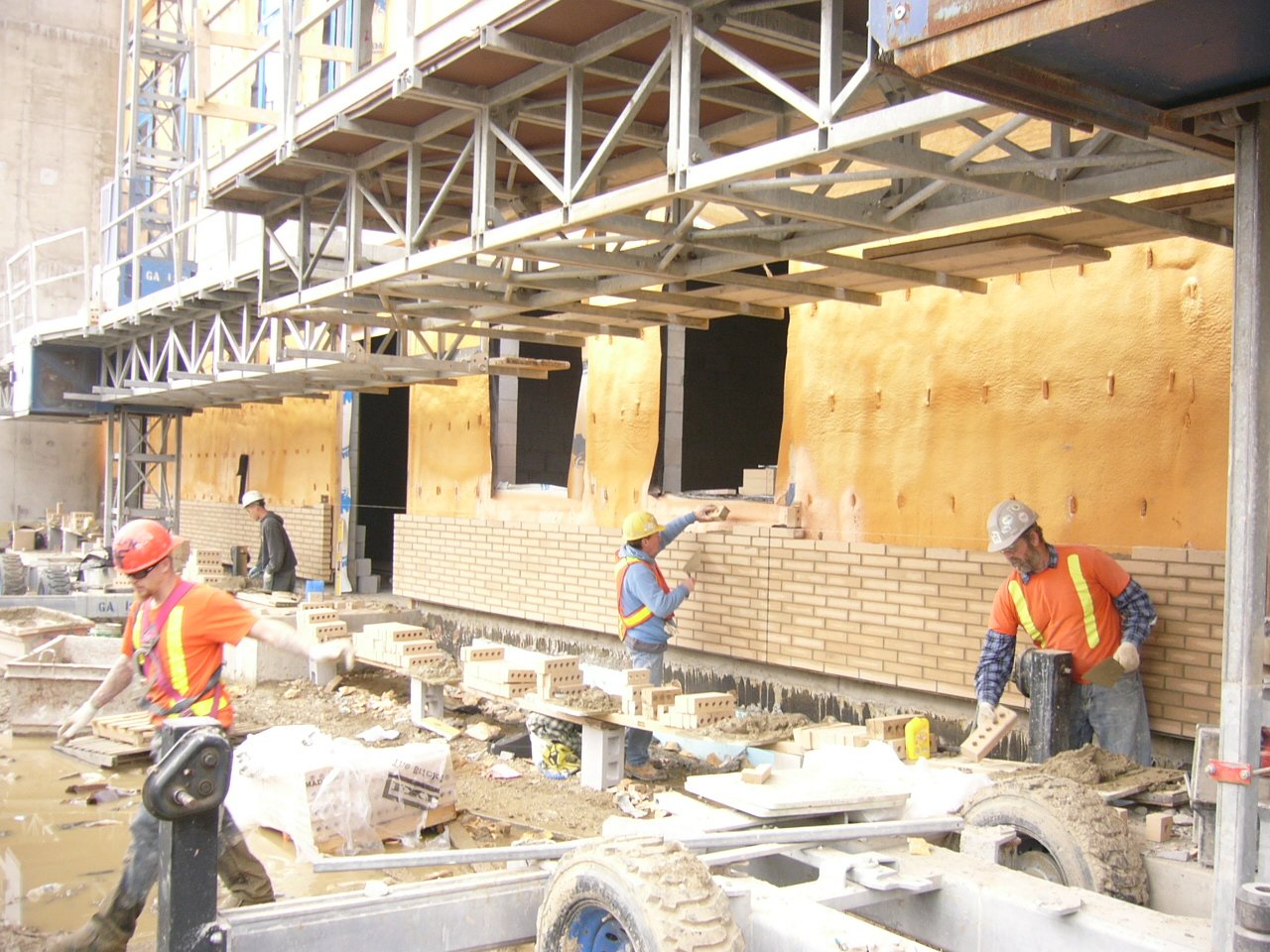 Construction workers building a brick wall on a construction site, with scaffolding above and various construction materials around.