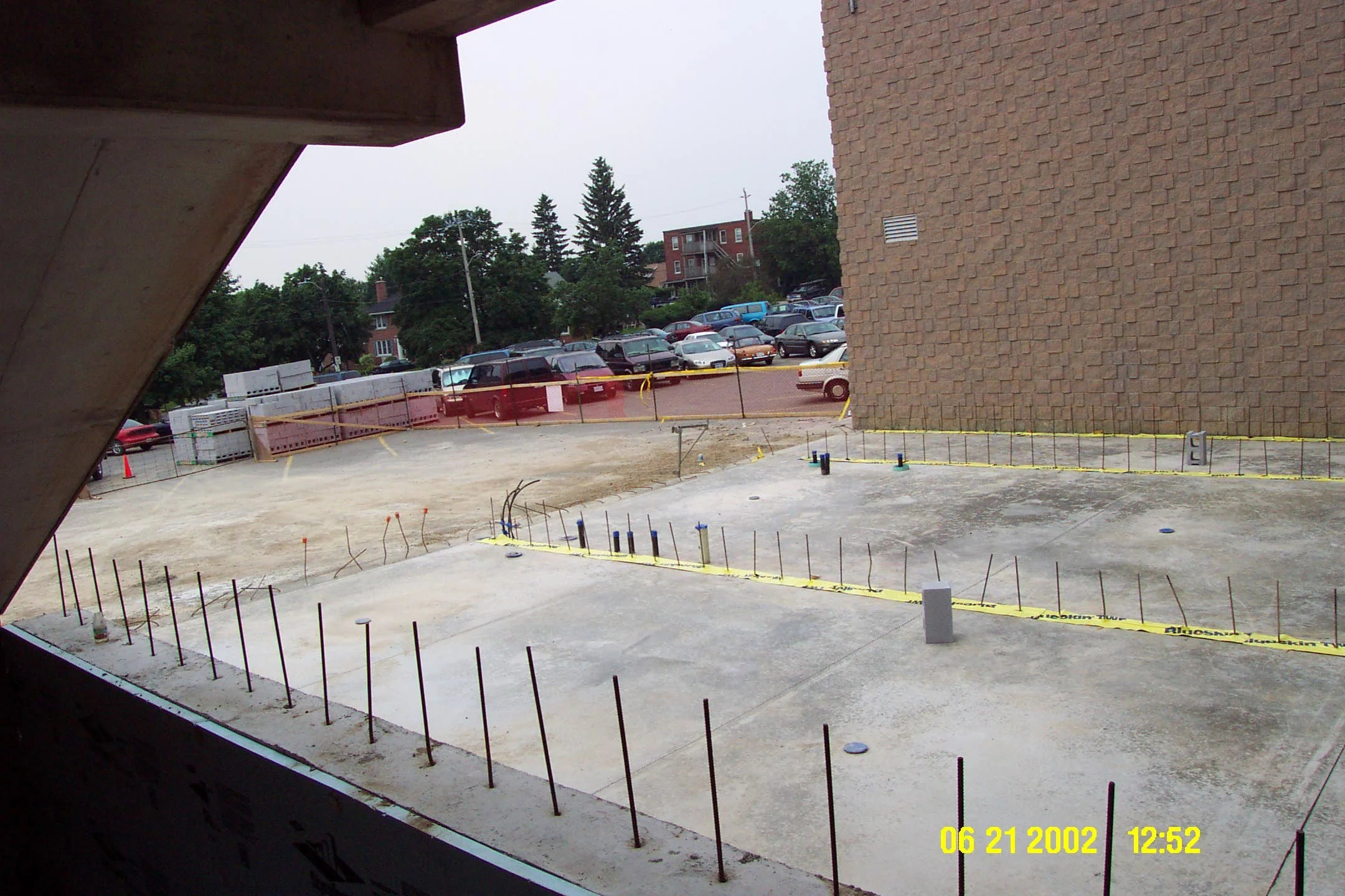 View of a construction site with concrete foundation, exposed rebar, and yellow caution tape, adjacent to a brick building with parked cars and trees in the background.