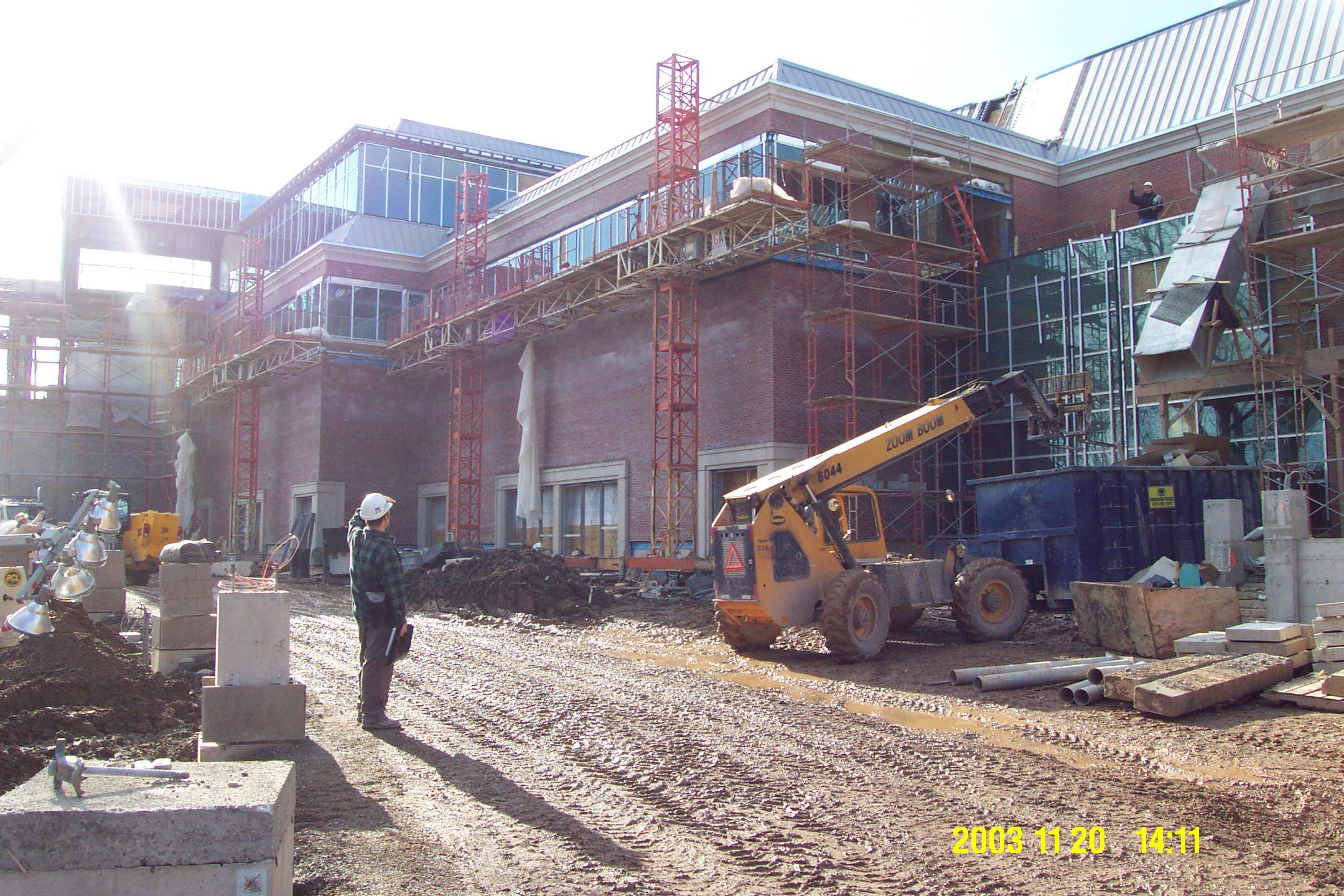 Construction site with a multi-story building under construction, scaffolding, and workers. A person in a white helmet stands on a dirt ground, and a yellow telescopic forklift is moving materials. Building materials and equipment are scattered on th