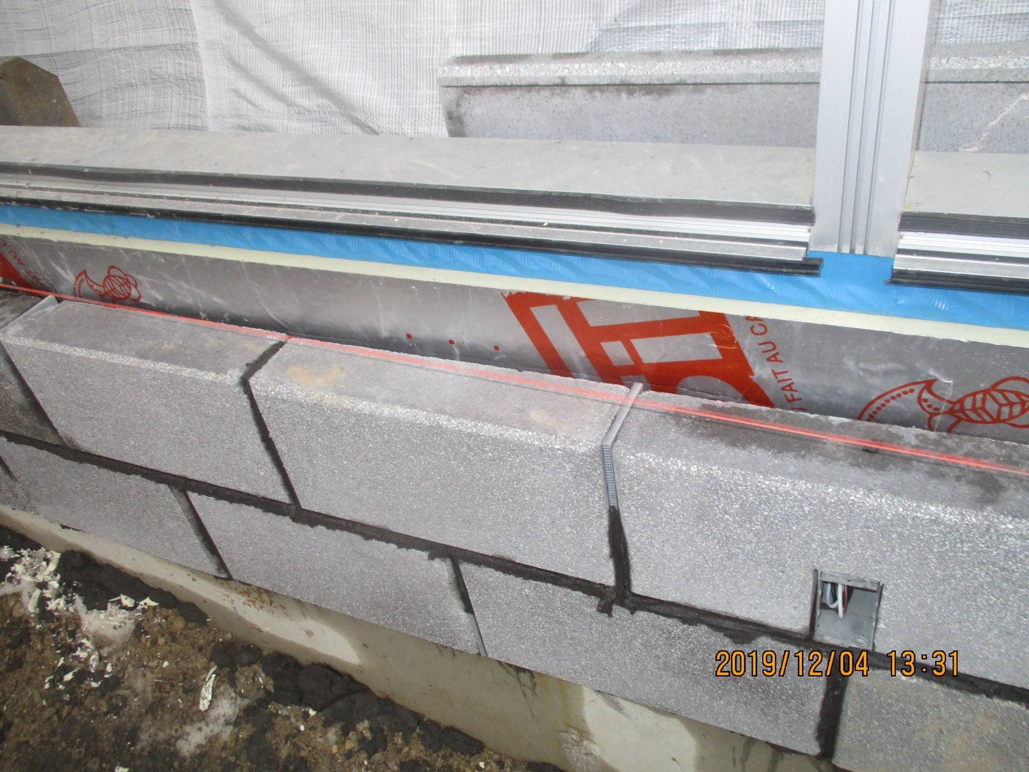 Close-up of a construction site showing a new cinder block wall with a metal window frame and weatherproofing barrier above it. The ground below is dirt with debris.