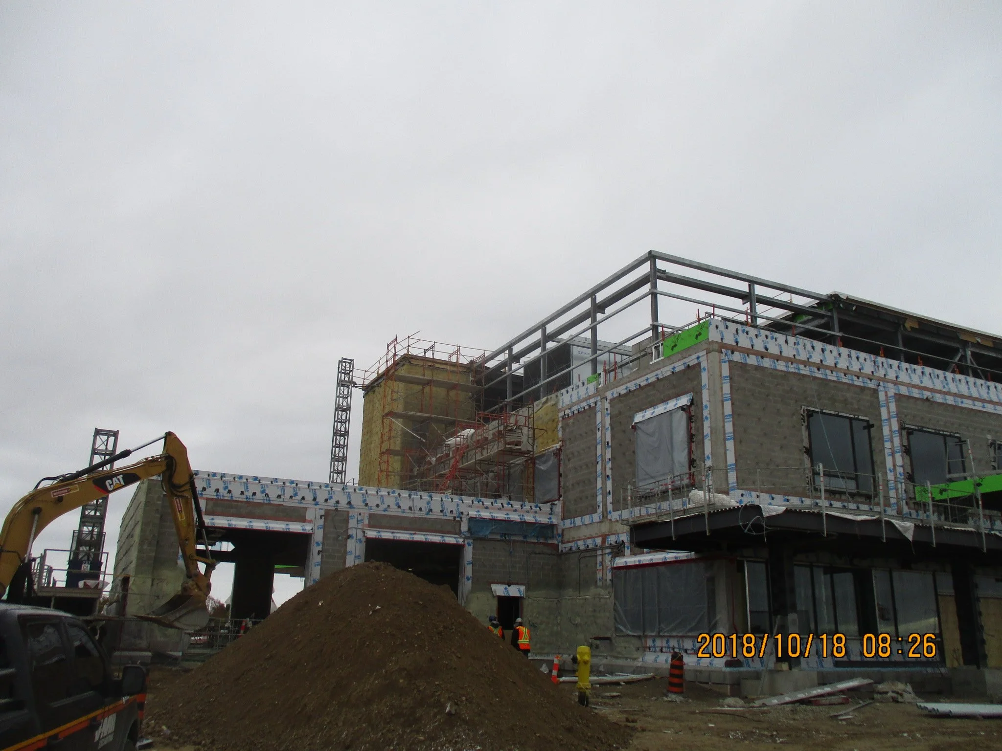 Construction site with a partially built multi-story building, scaffolding, construction workers, and an excavator with a large pile of dirt in the foreground.