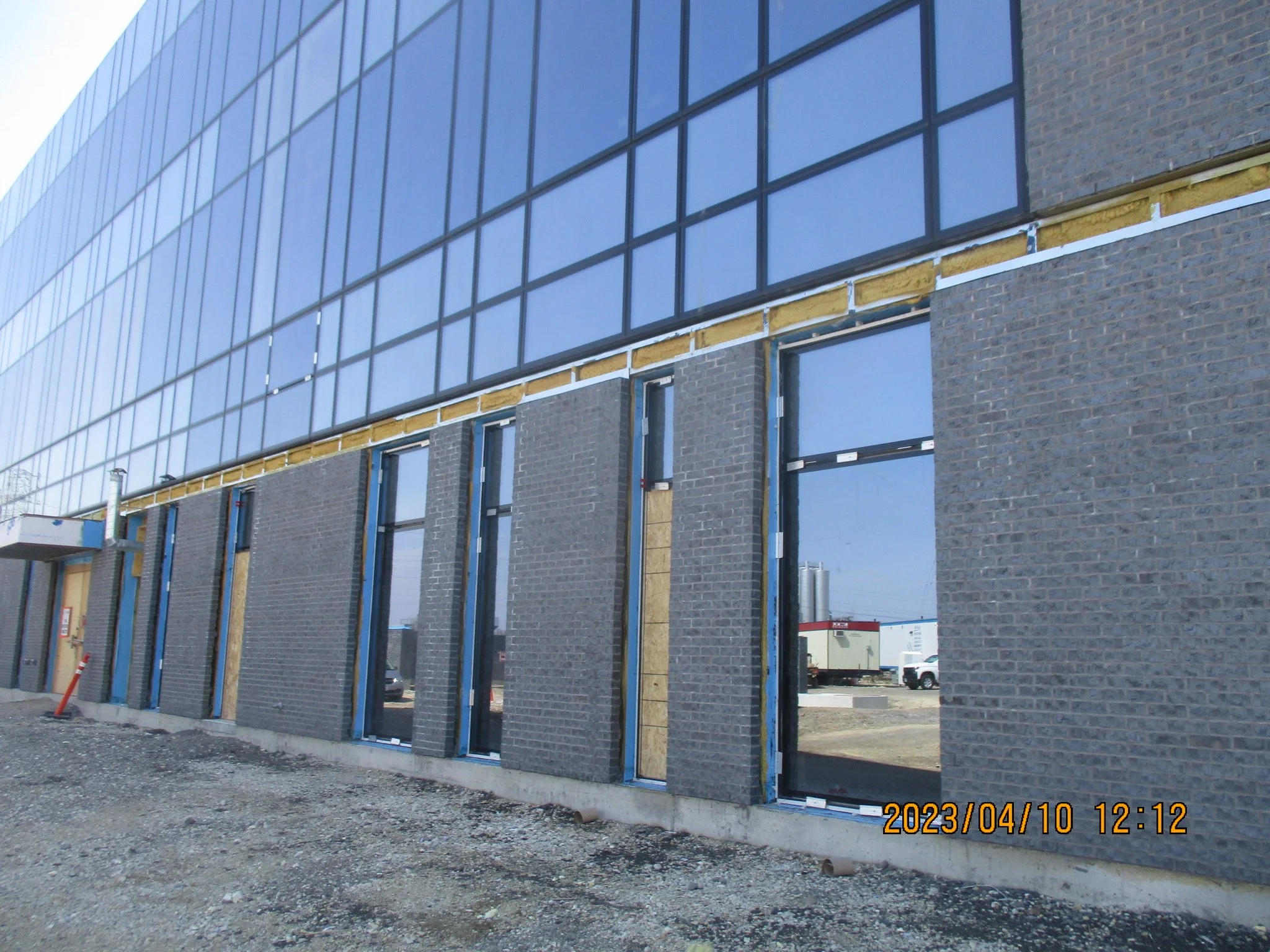 Building under construction with brick and glass exterior, doors and windows being installed, construction materials on site, and construction equipment visible.