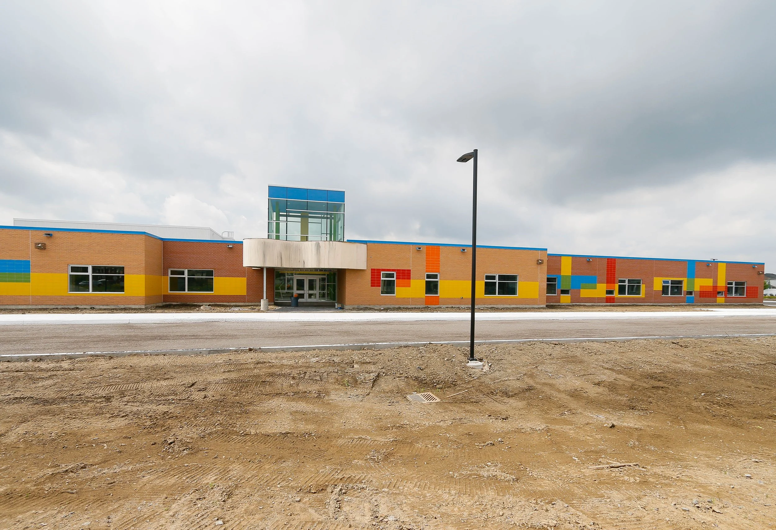 A colorful modern building with a brick facade, decorated with horizontal yellow, red, and blue stripes, and geometric patterns. The building has large windows and a glass entrance with a small overhang, set against a cloudy sky. In the foreground, t