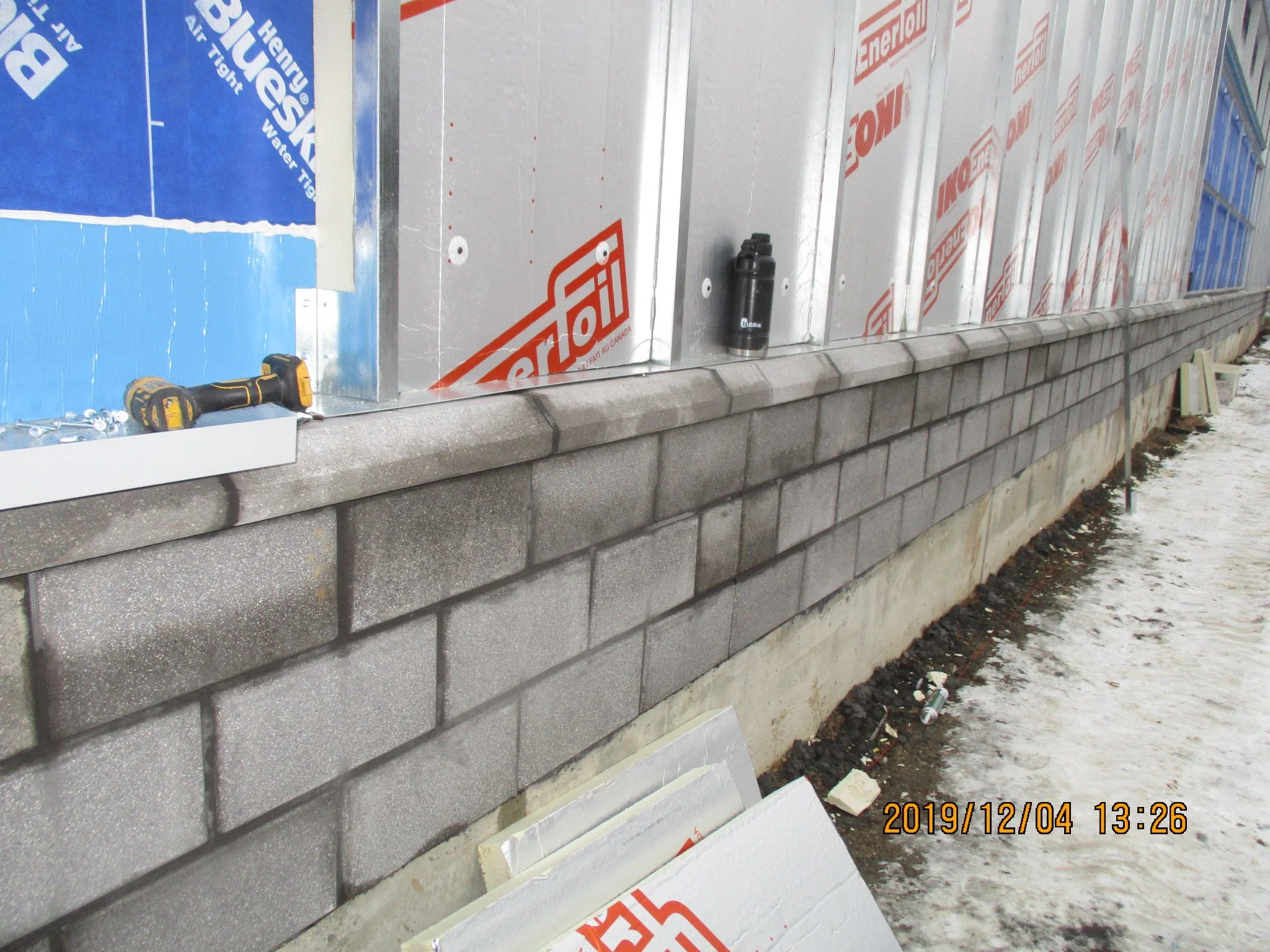 Construction site showing a partially built brick wall with construction materials, tools, and insulation panels nearby, snow on the ground.