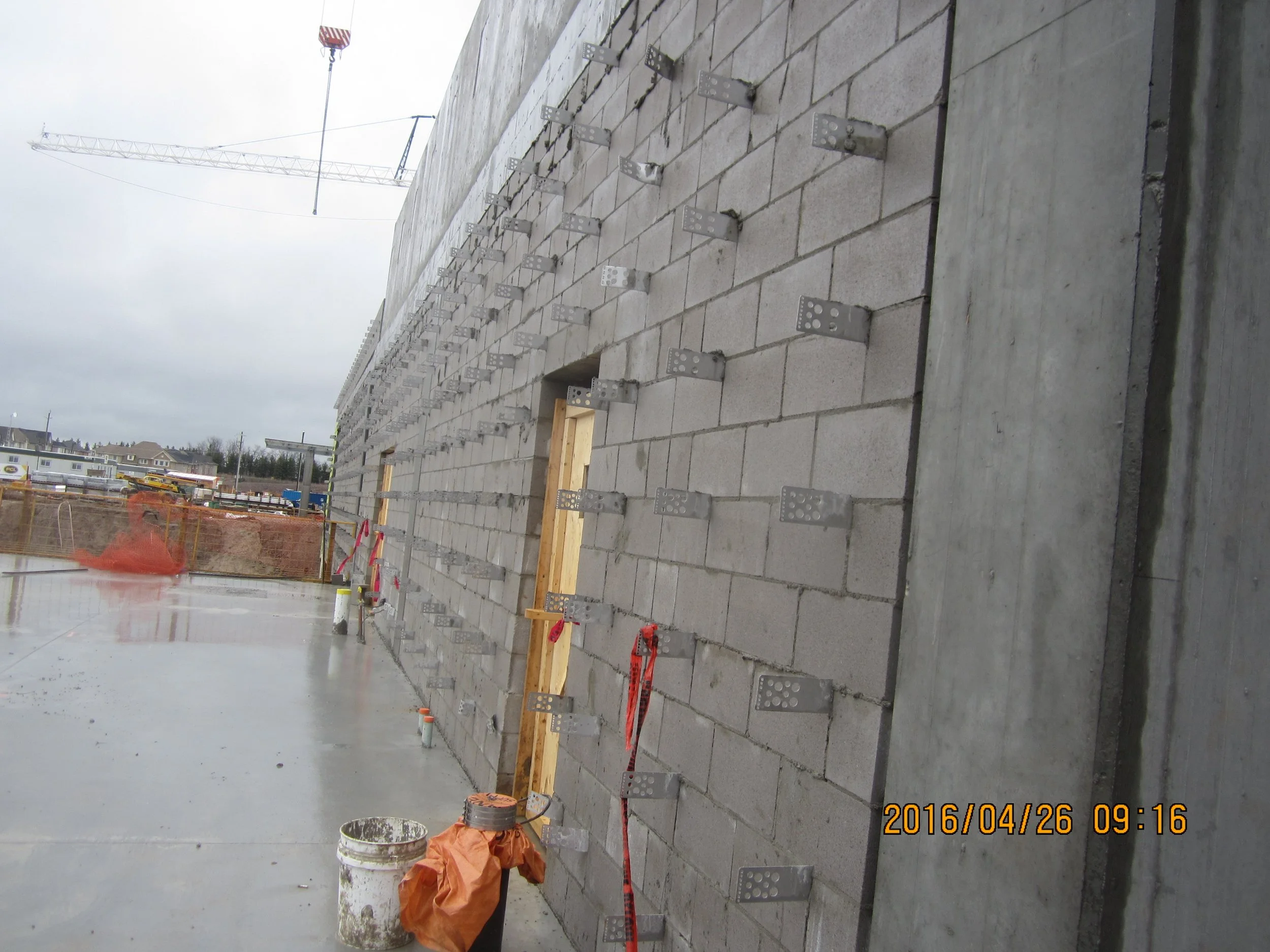Construction site showing a building wall with metal fasteners attached to concrete blocks, wooden frame, and construction materials on wet ground.