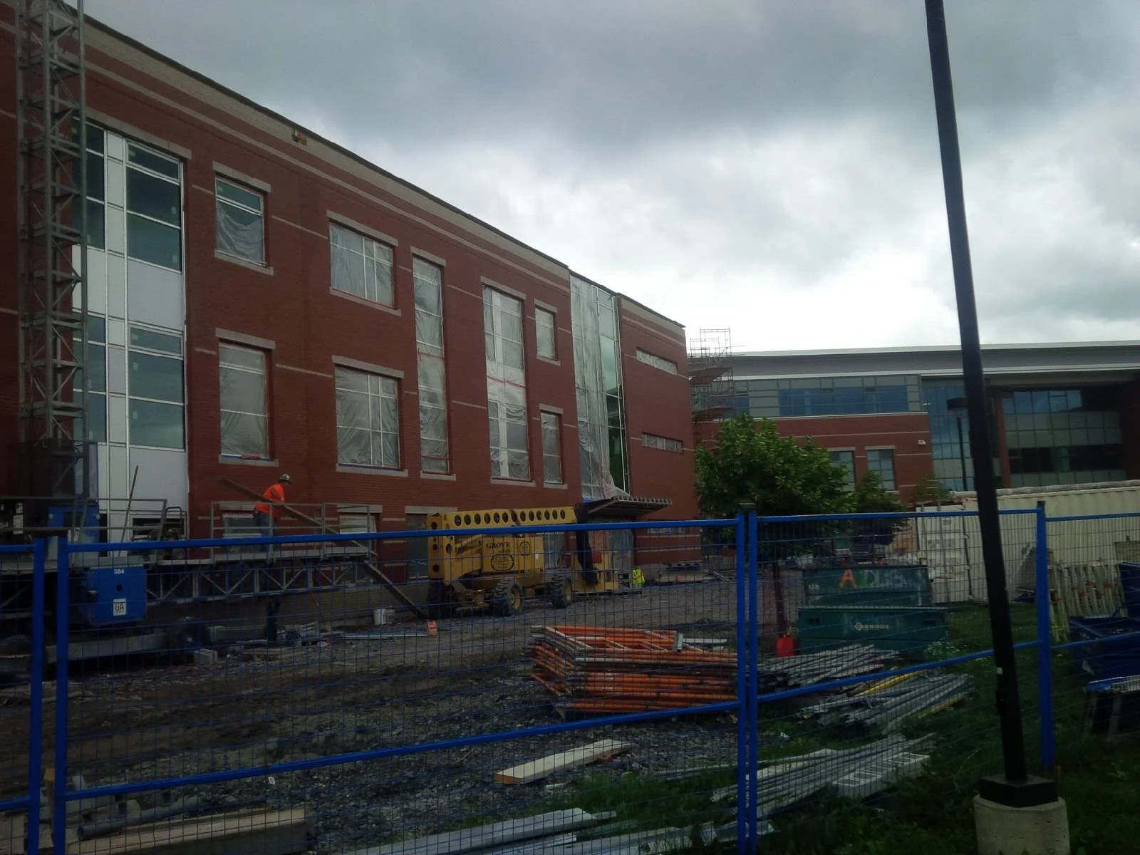 Construction site with workers, scaffolding, and building materials in front of a modern brick and glass building, under a cloudy sky.