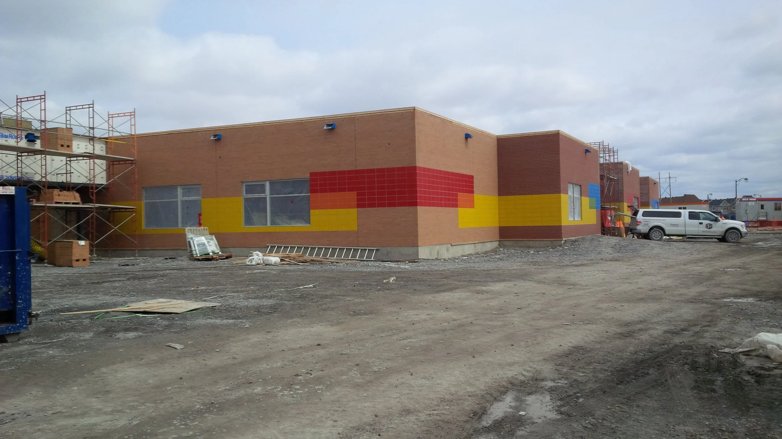 Construction site of a building with colorful brick facade, scaffolding, construction materials, and a white pickup truck parked nearby on a dirt lot under cloudy sky.
