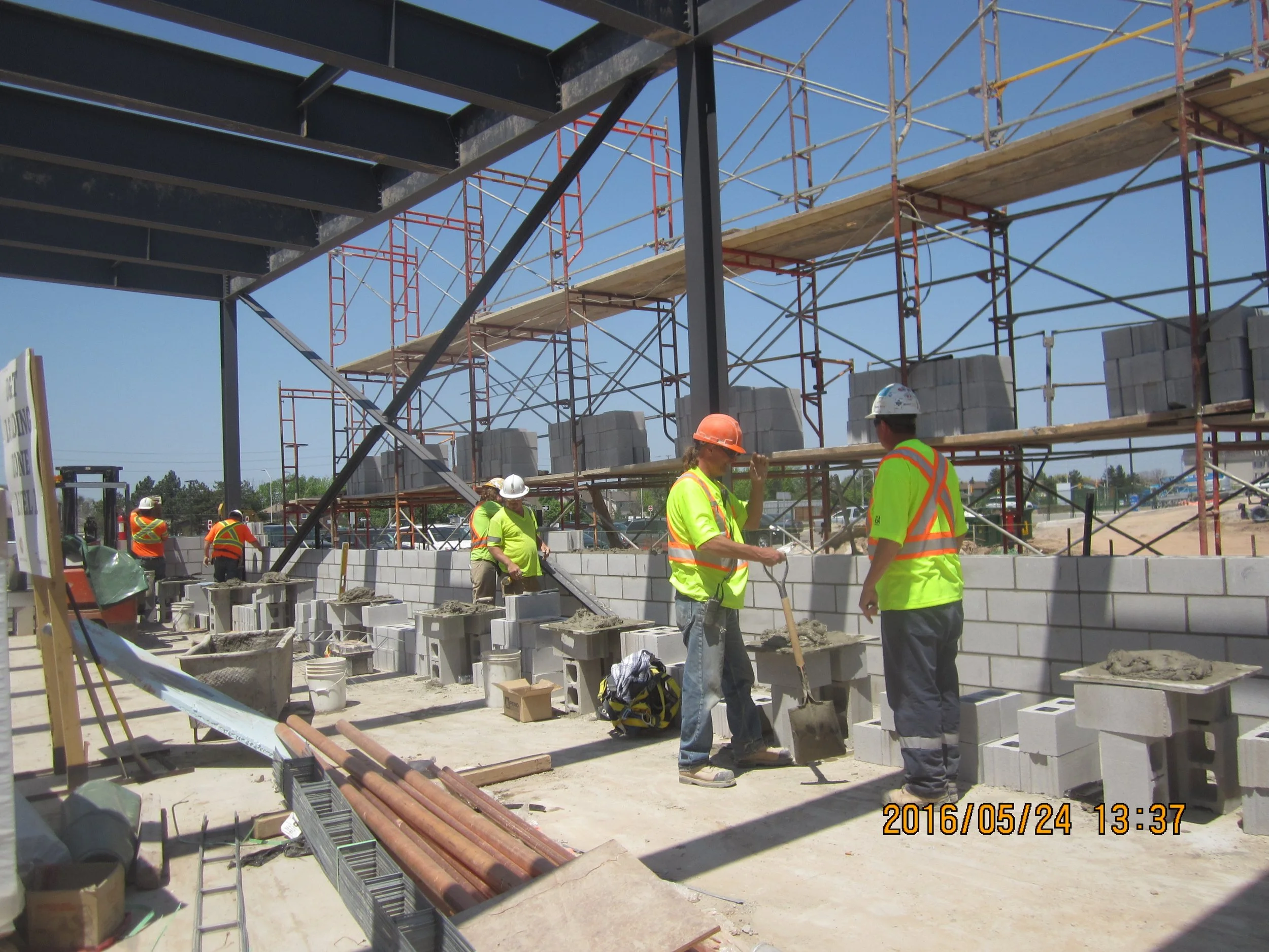 Construction workers in safety vests and helmets building a brick wall at a construction site with metal scaffolding and supplies around.