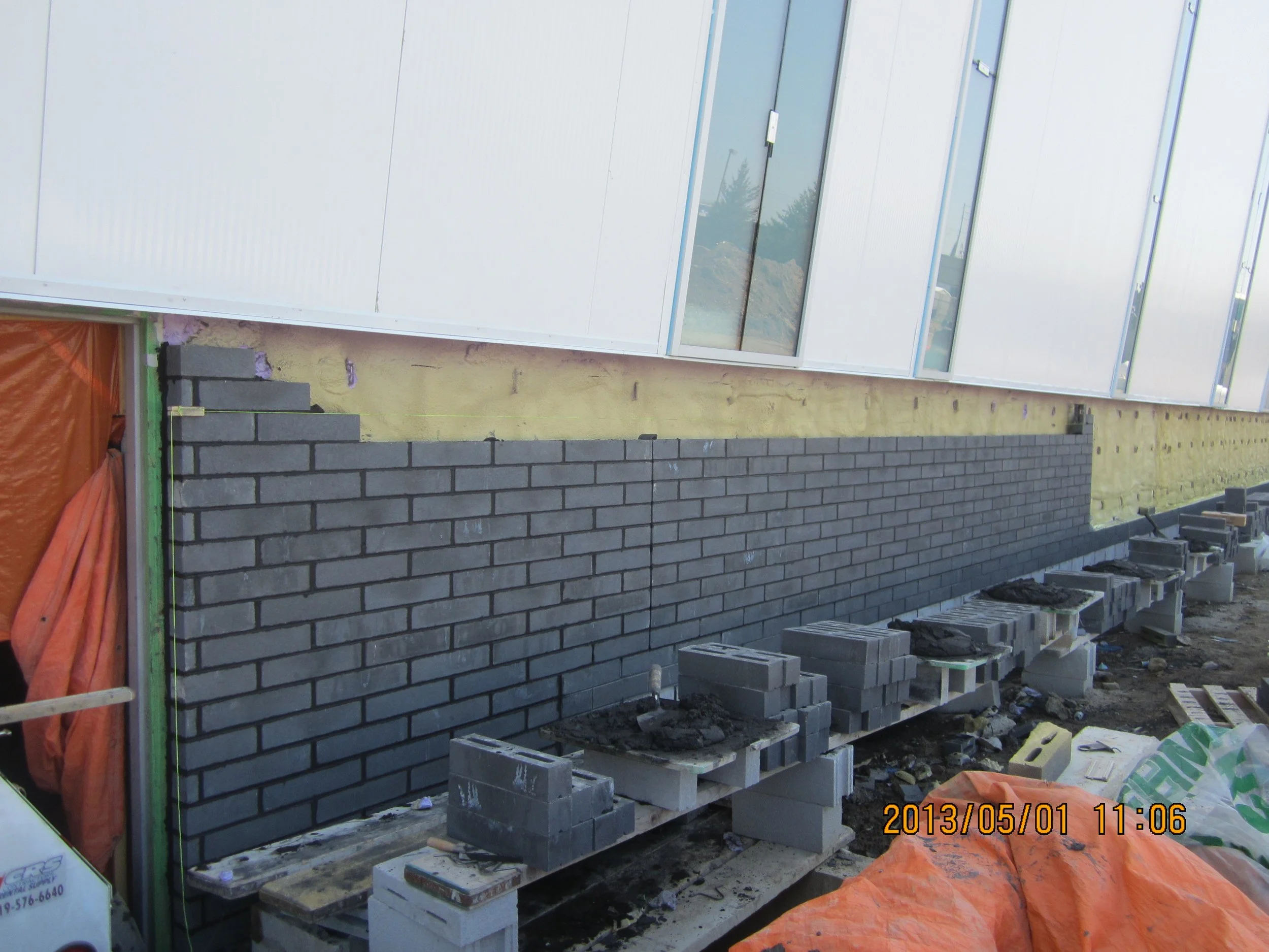 A black brick wall being constructed outside a building with white panel siding and large windows, with construction materials and pallets nearby.