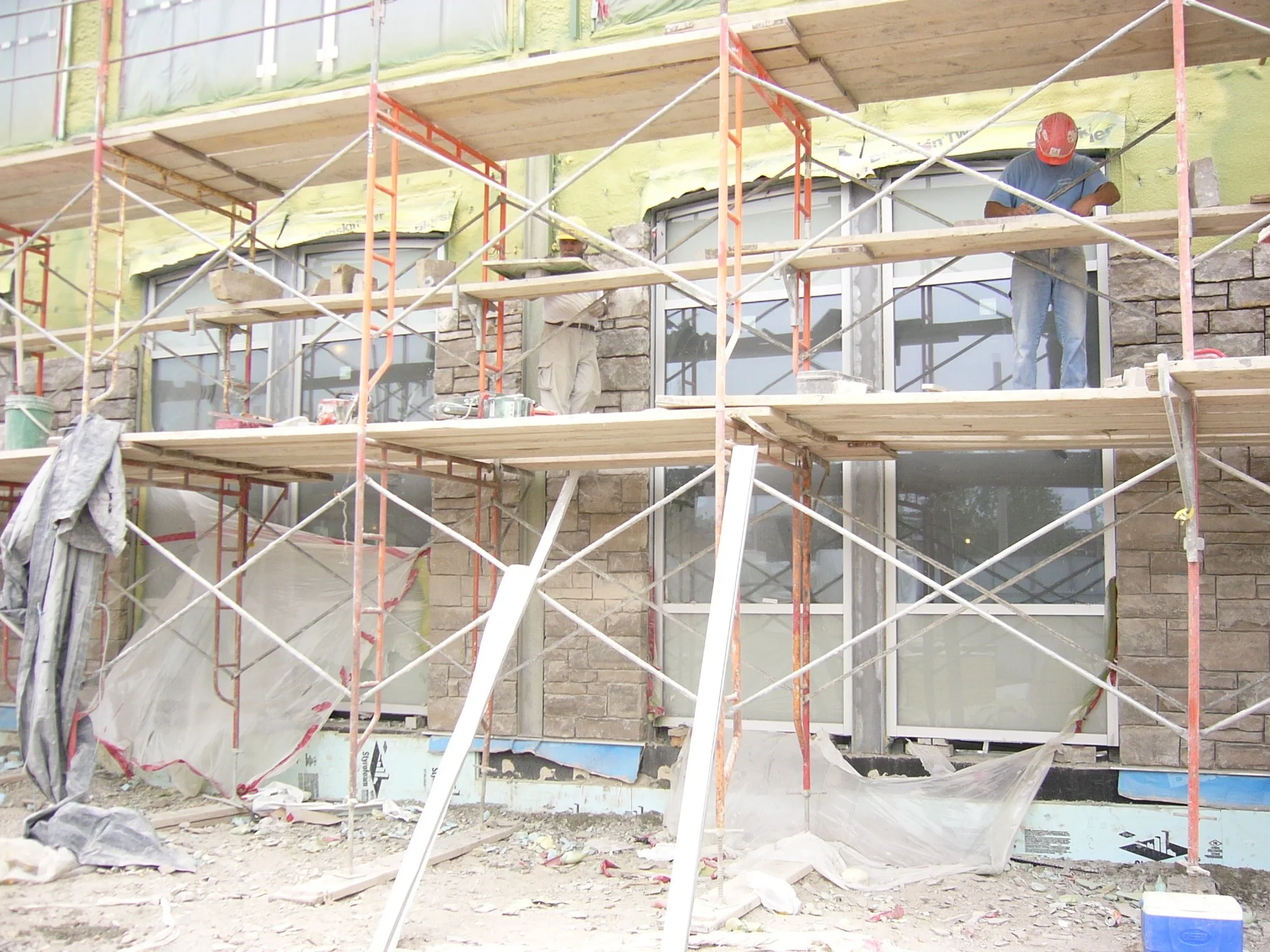 Construction workers on scaffolding installing stone veneer on a building facade with large windows.