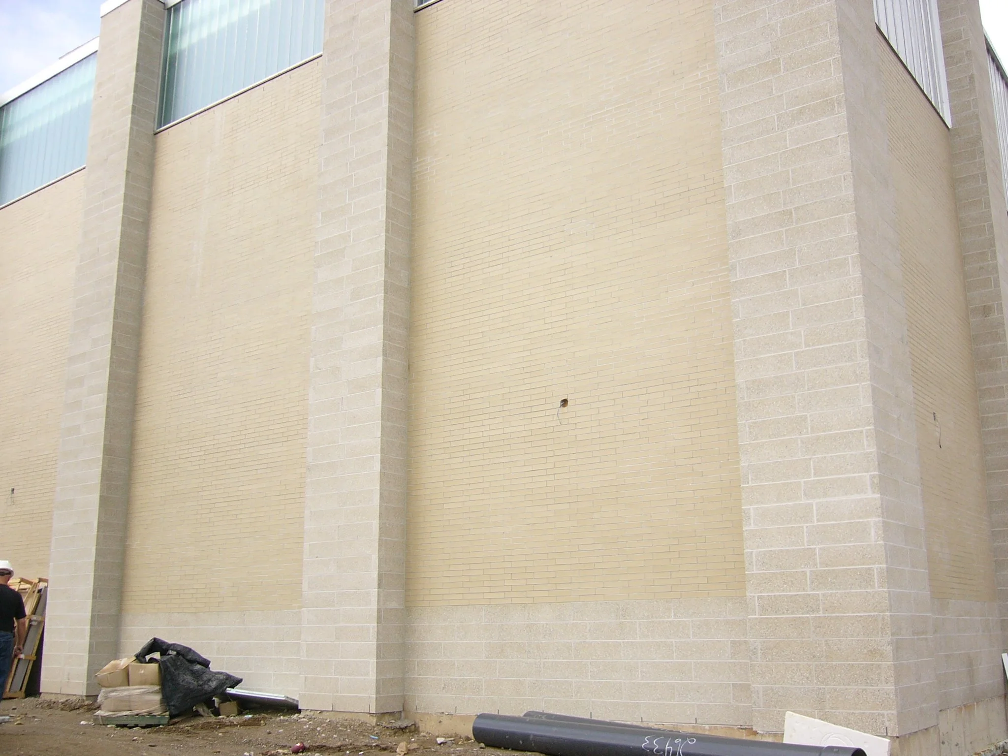 Exterior view of a building with a beige brick facade and large windows at the top, construction materials and debris at the bottom.