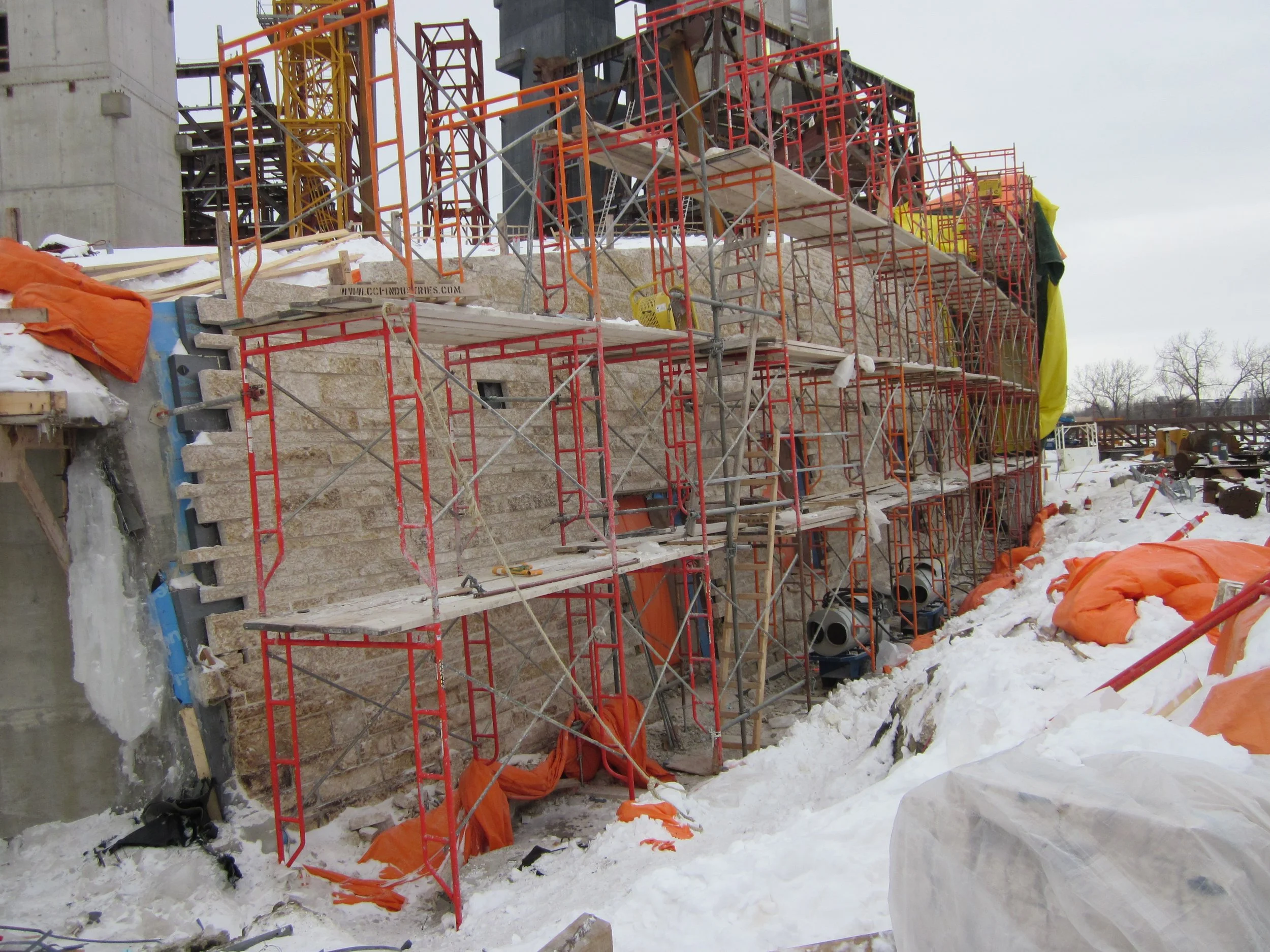 Construction site with scaffolding and stone wall, snowy ground with orange and white tarps, and construction equipment.