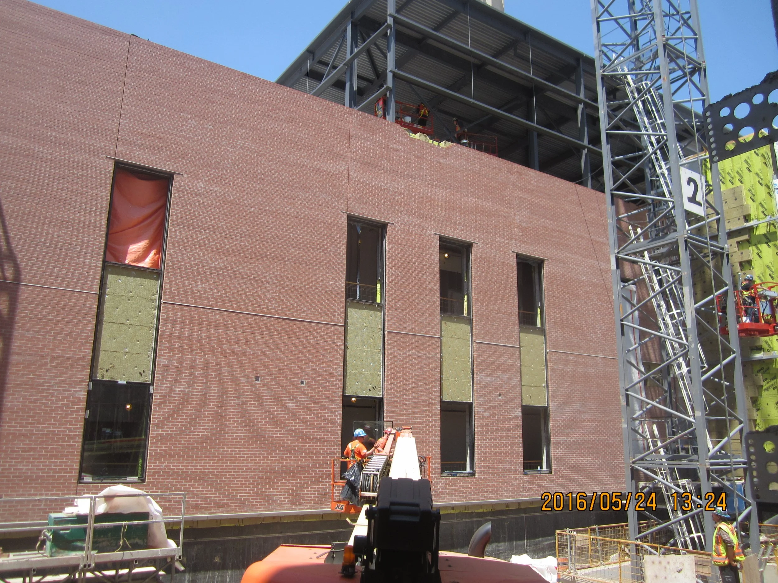 Construction workers installing window frames on a brick building under construction, with a large steel scaffolding and a crane.