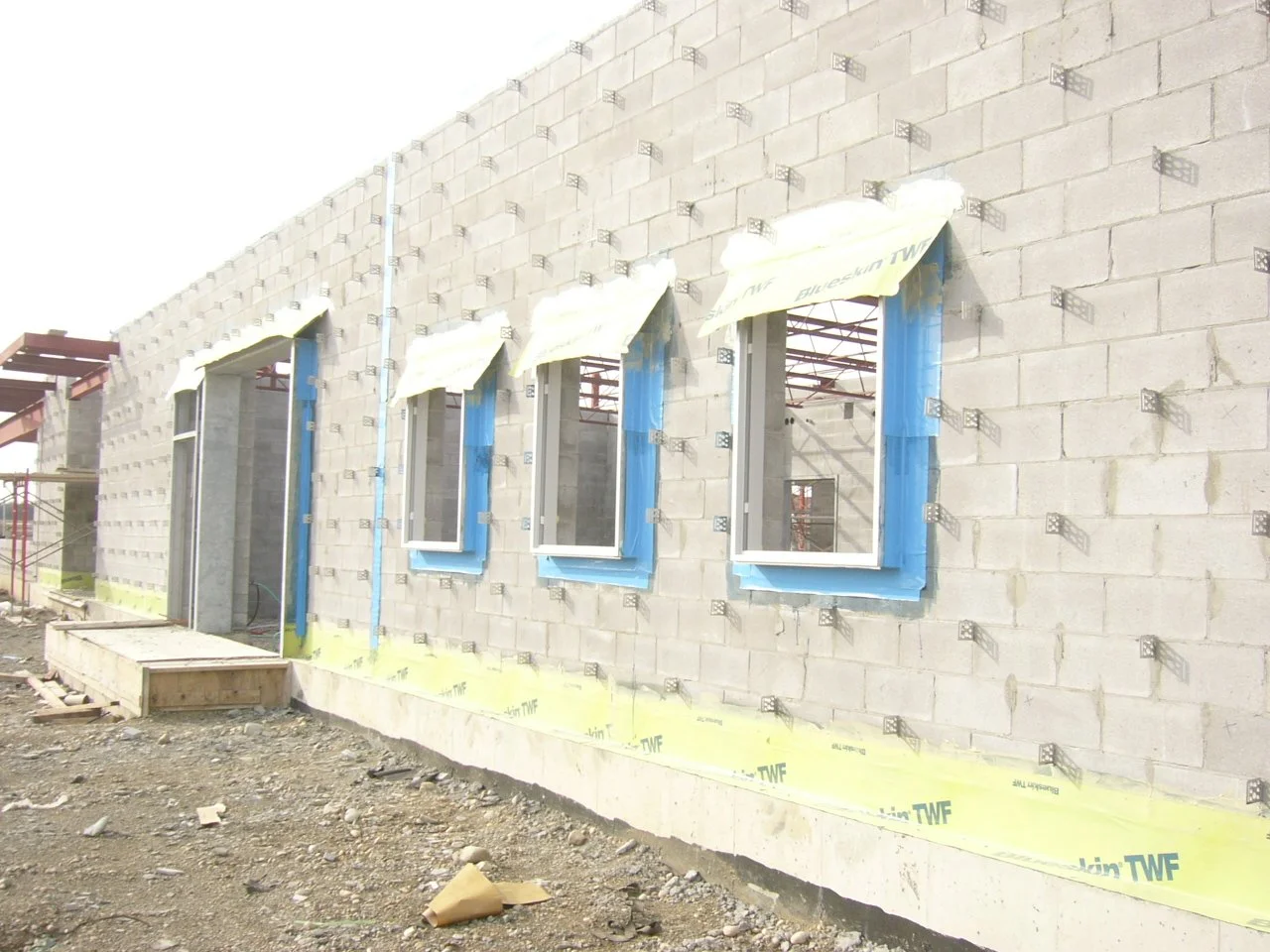 Under construction building with unfinished brick walls and three window openings with blue window frames, construction material and scaffolding present.
