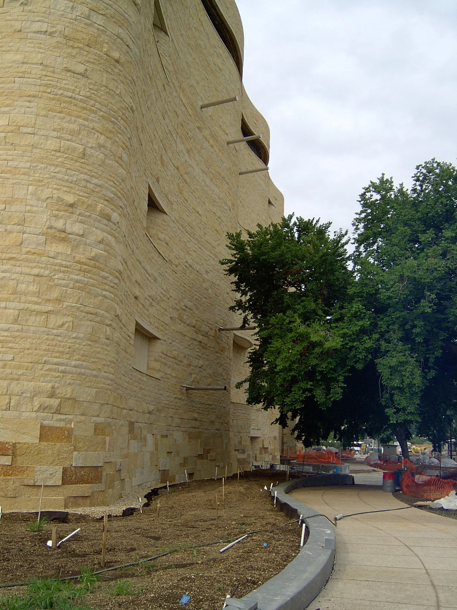 Under construction sidewalk along a large, curved stone building with small open windows, with trees and construction materials nearby.
