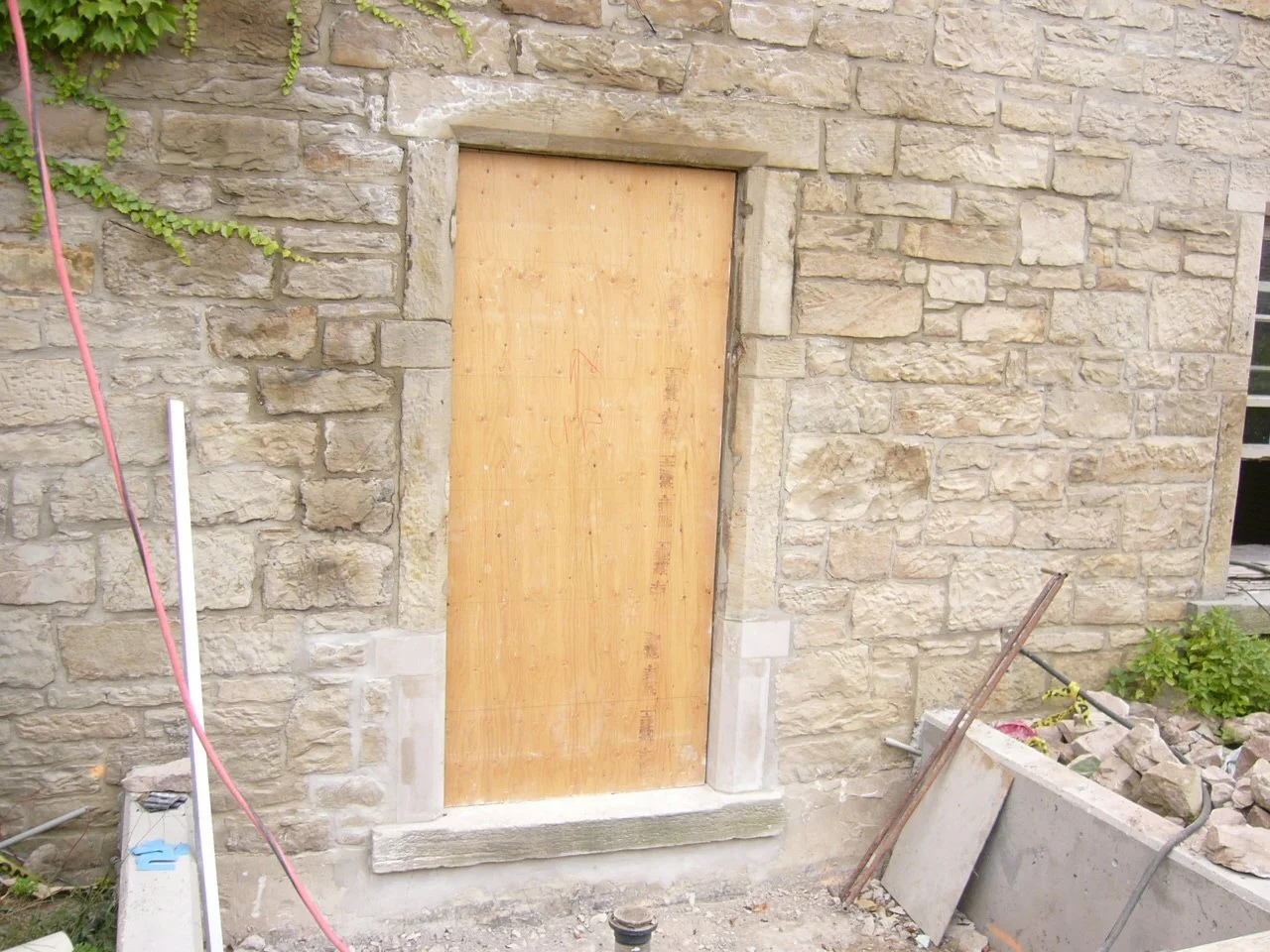 A stone wall with a wooden boarded-up window, construction tools and debris in the foreground.