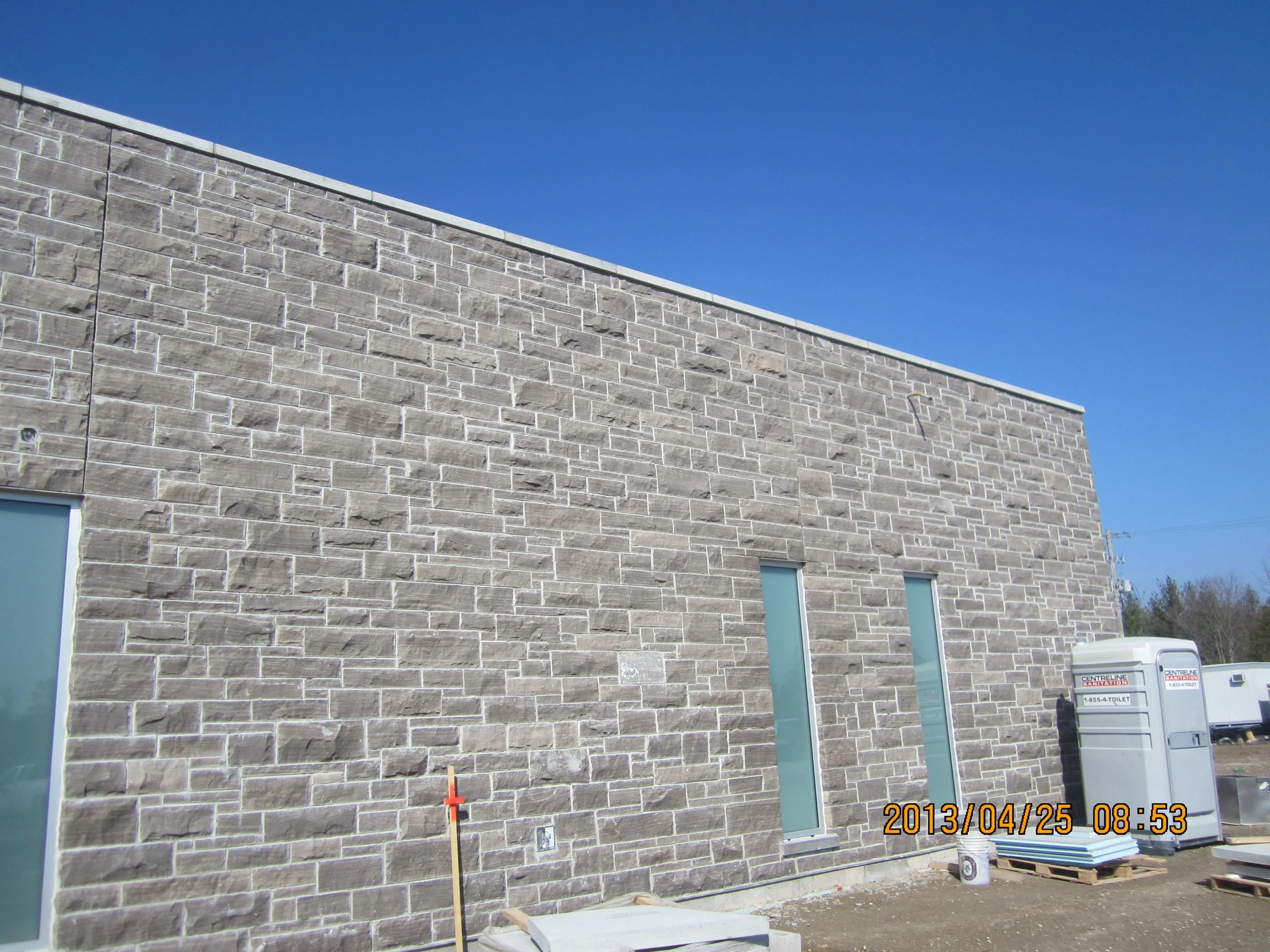 Exterior brick wall of a building under construction with two tall, narrow windows, construction equipment, and a portable toilet, against a bright blue sky.