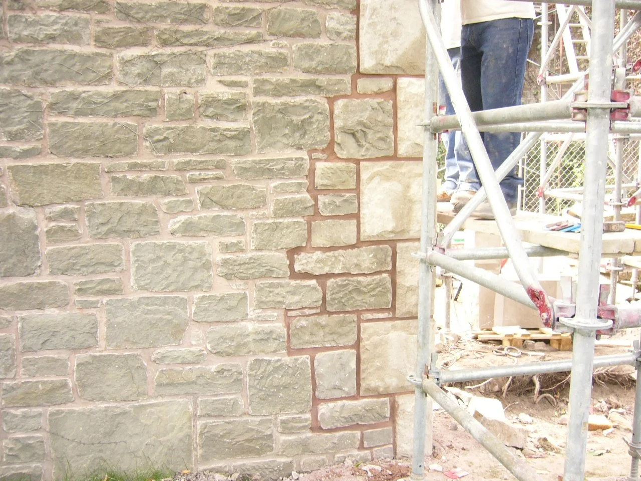 Close-up of a stone brick wall under construction with scaffolding and worker's legs visible.