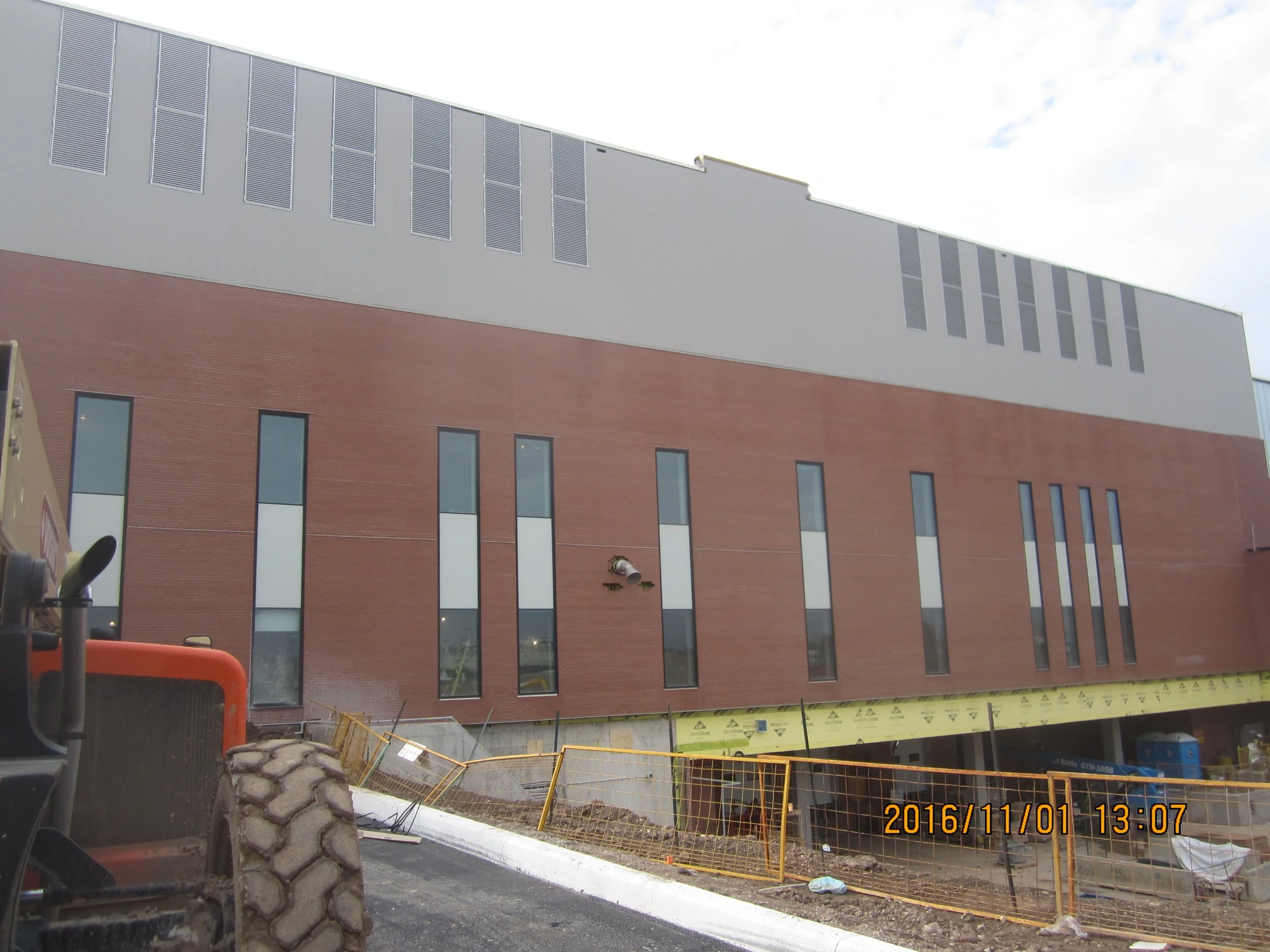 Construction site with a building having a red brick lower section and a gray upper section, with several narrow vertical windows and some construction barriers and equipment visible in the foreground.