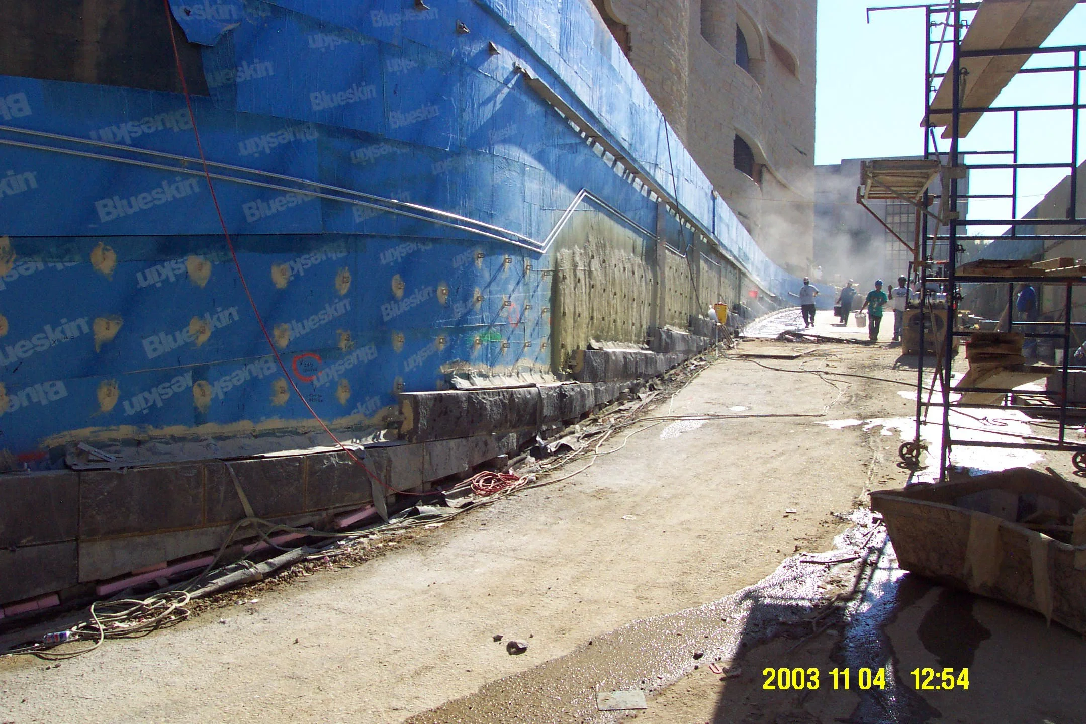 Construction site with workers, scaffolding, and building materials; blue weather-resistant sheeting on the wall, dirt ground with some pipes and wires, sunlight, and steam or smoke in the background.
