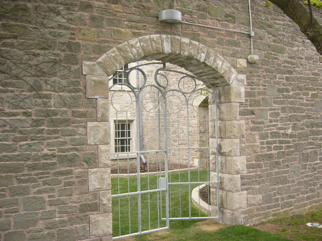 A metal gate with circular and straight patterns in front of a stone arched doorway on a brick wall, leading to a garden with grass and a stone walkway.