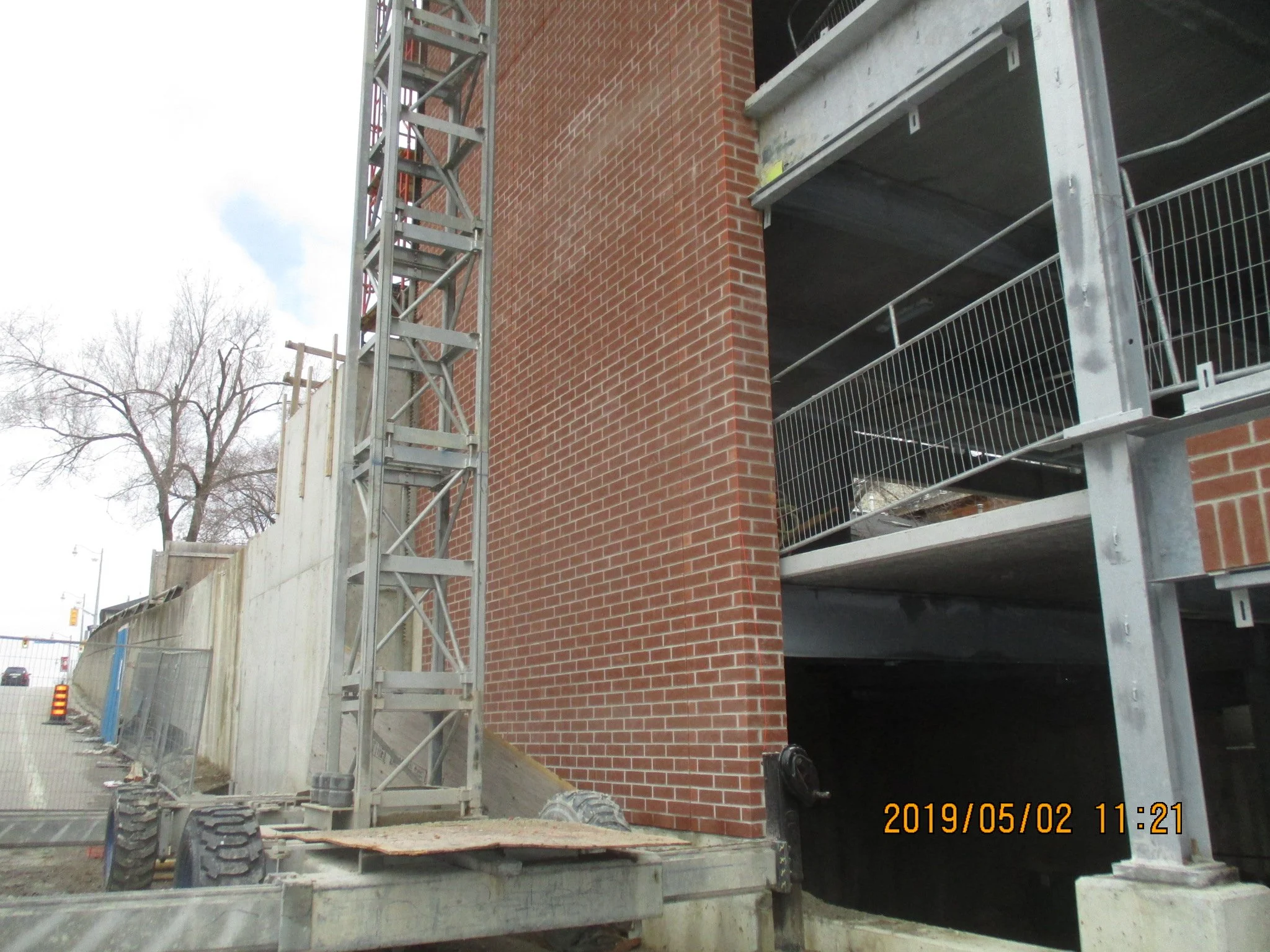 Construction site with metal scaffolding and brick building wall, viewed from ground level, with trees and partly cloudy sky in background.